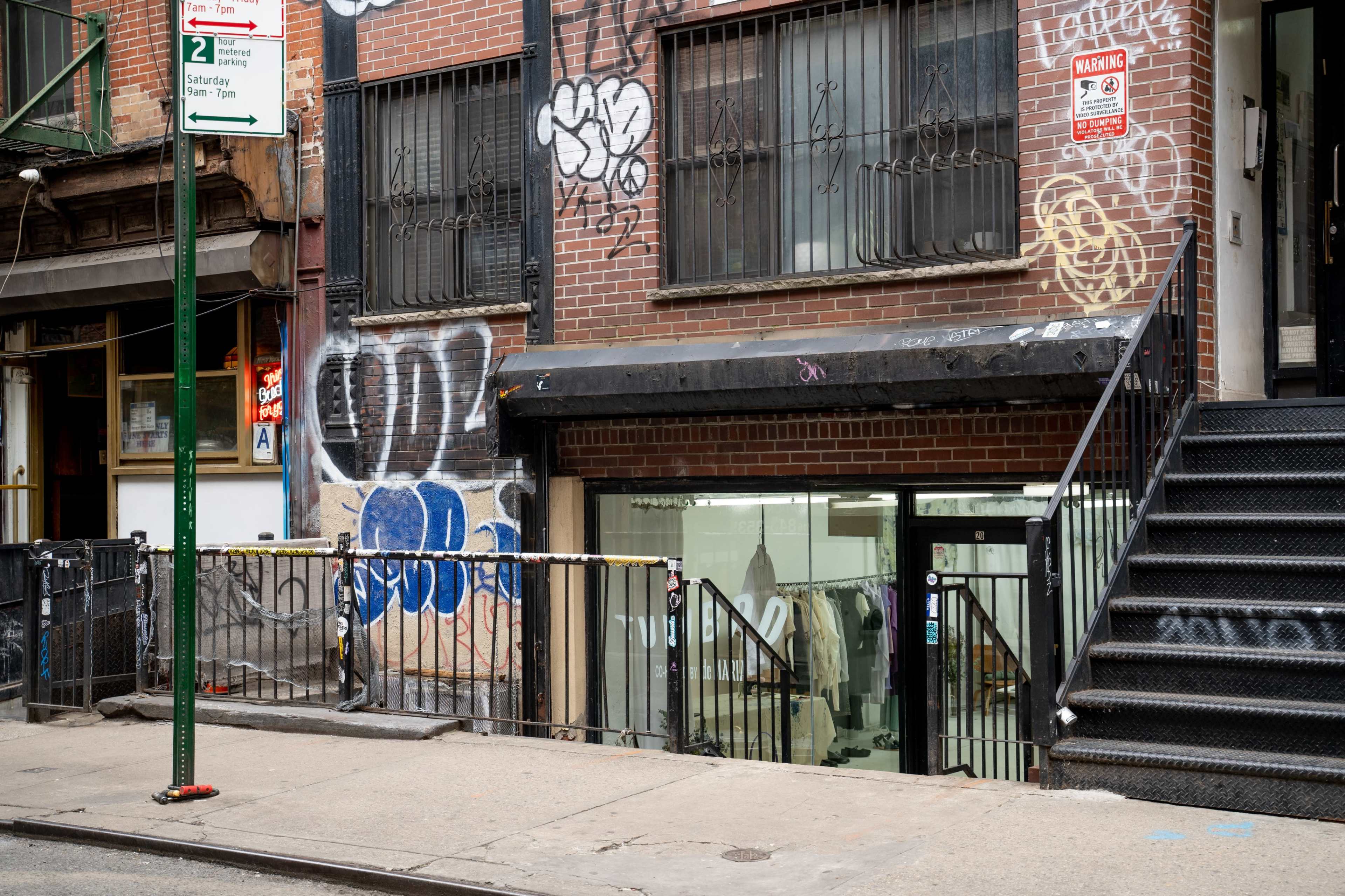 A brick building with graffiti on the exterior features a staircase leading up to a glass storefront entrance.