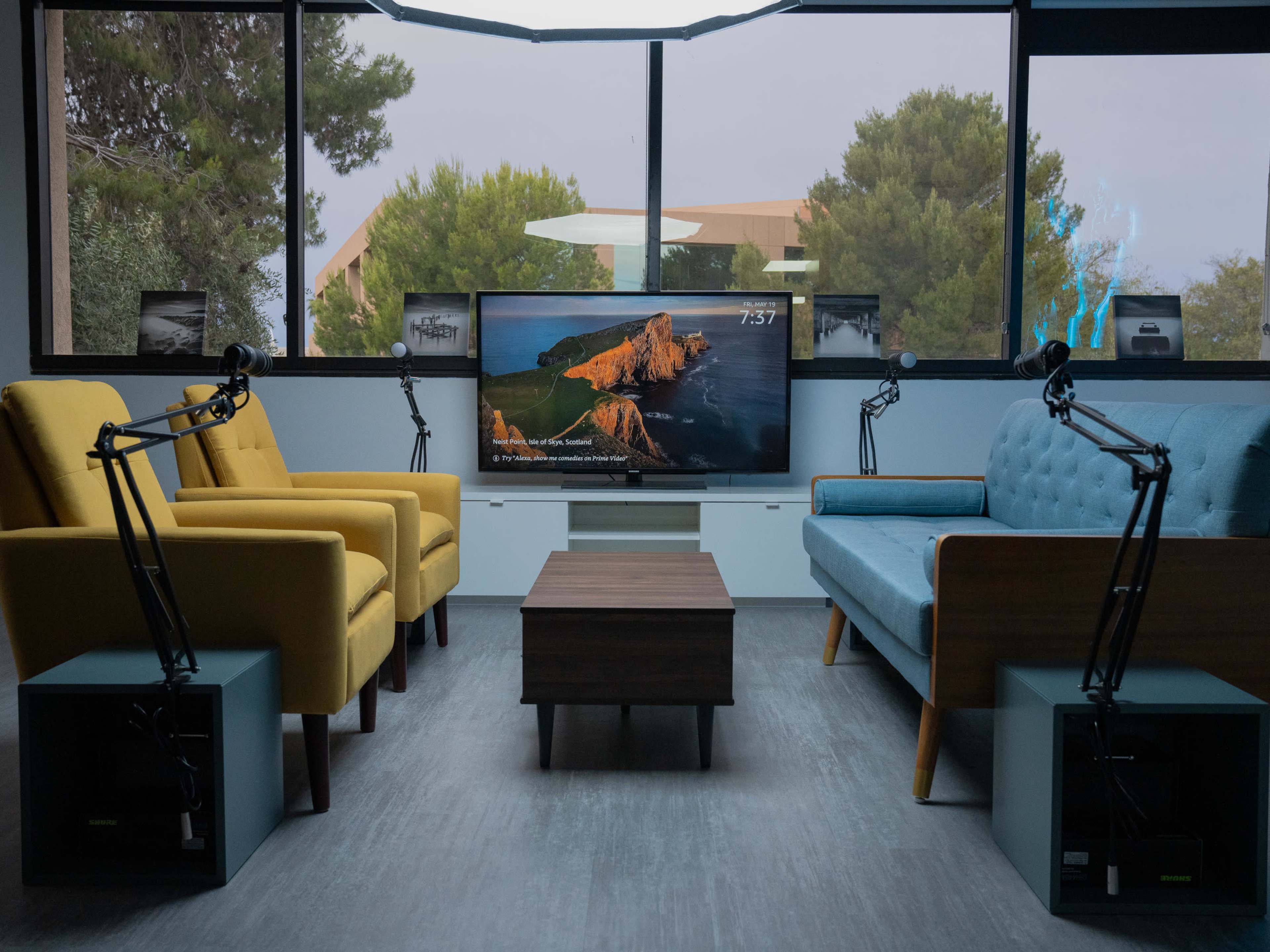 A modern living space features two armchairs, a blue couch, a coffee table, and a television mounted on the wall, all set against a large window with trees outside.