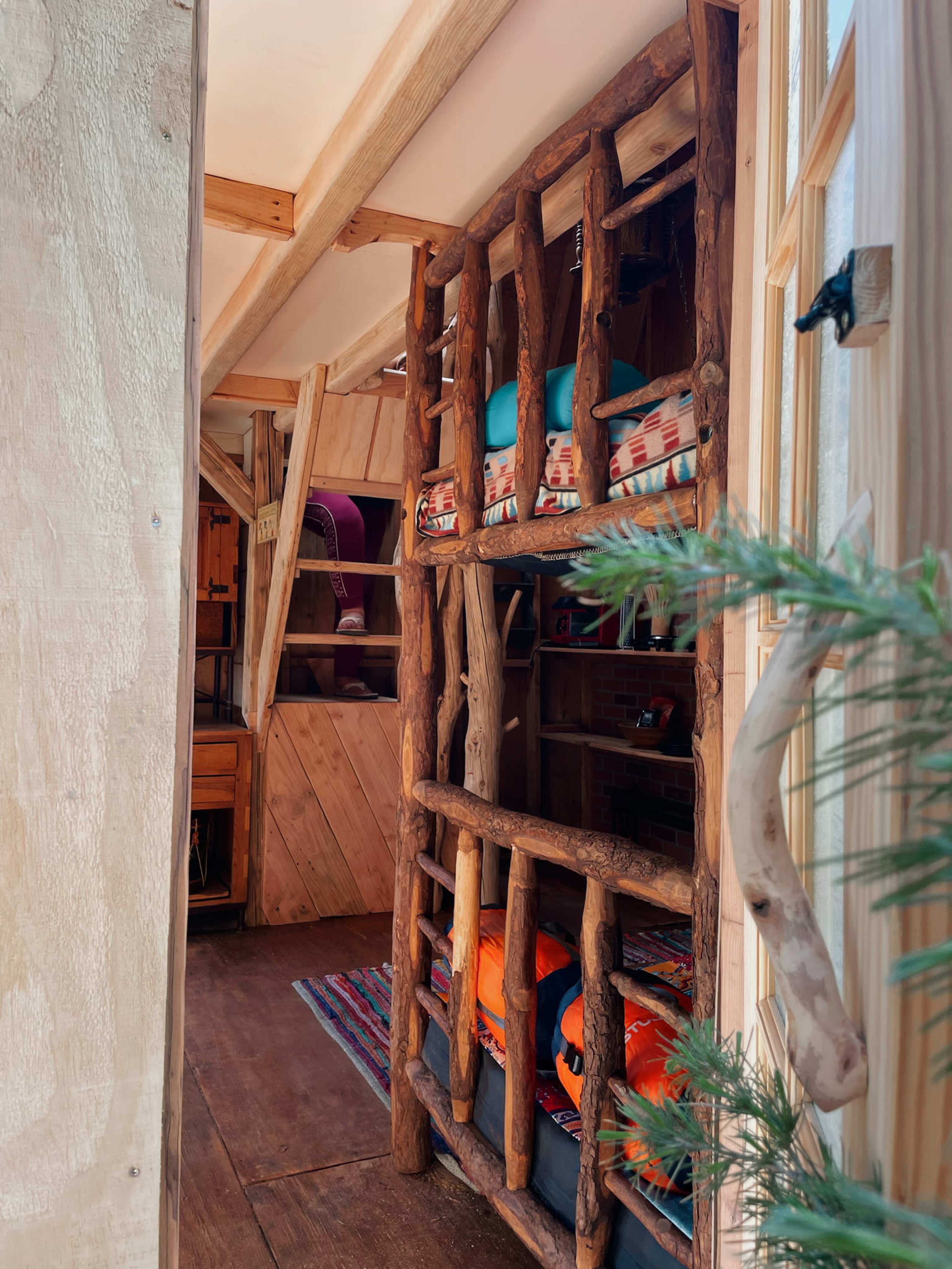 The interior of a cozy cabin features a wooden structure with bunks made of logs, colorful bedding, and a warm, inviting atmosphere.