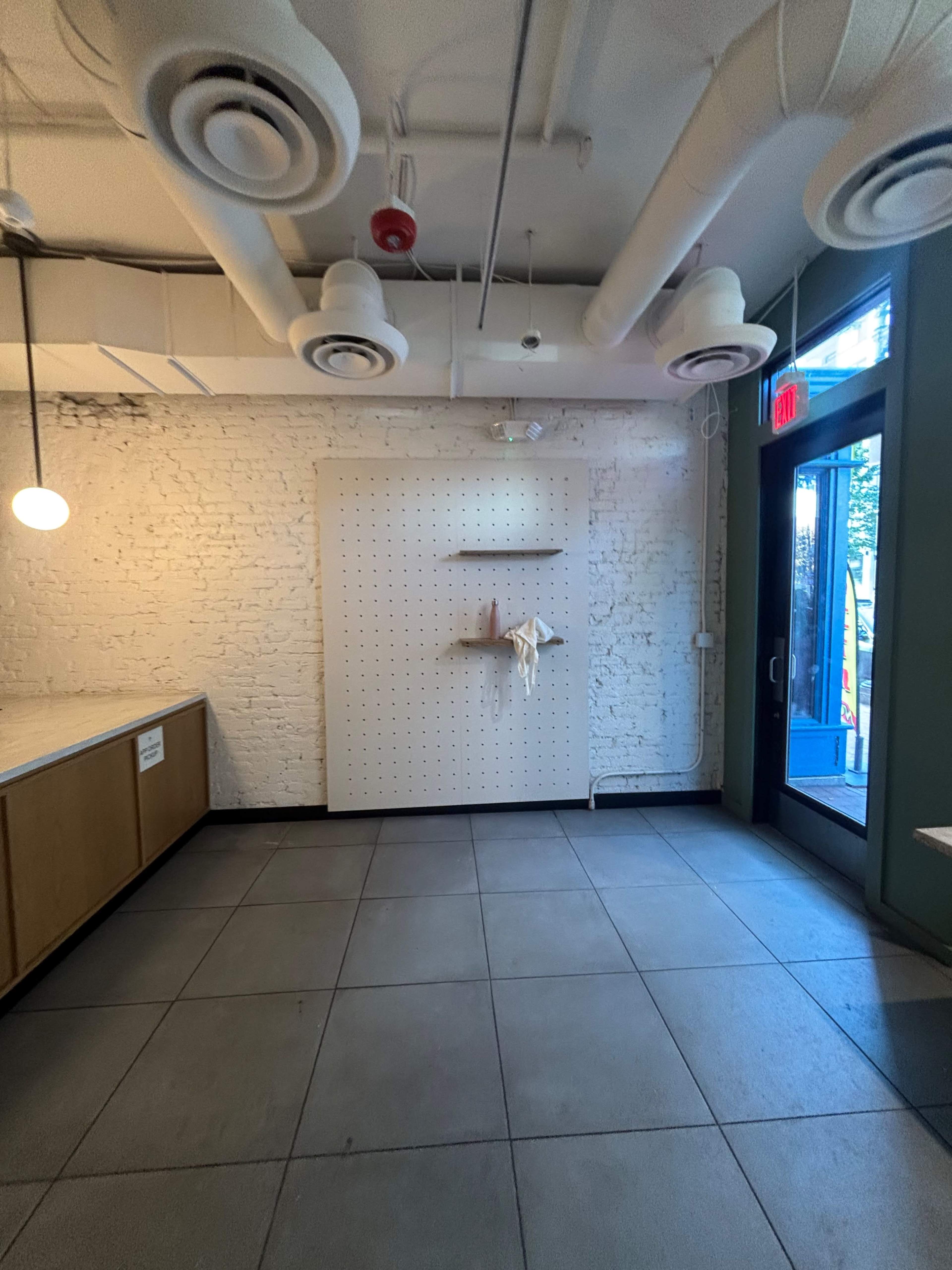 The image shows an interior space with exposed brick walls, a pegboard, and large windows leading to an outdoor area.