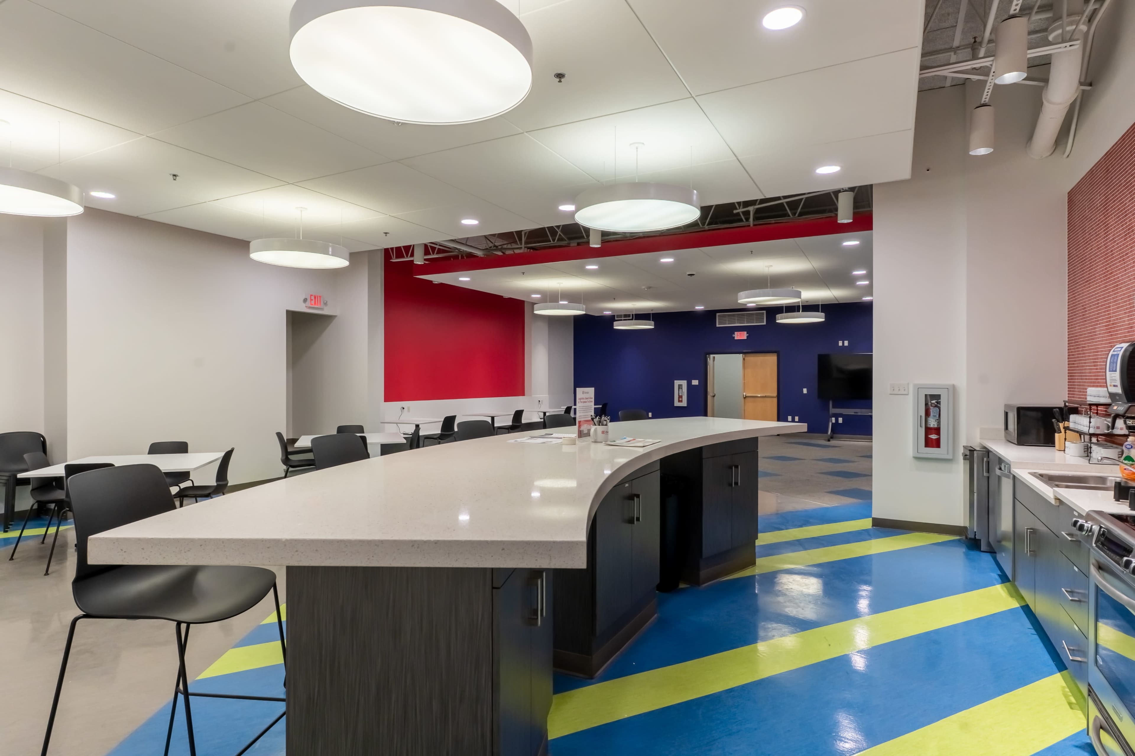 The image shows a modern break room with a large curved countertop, black chairs, and brightly colored flooring.