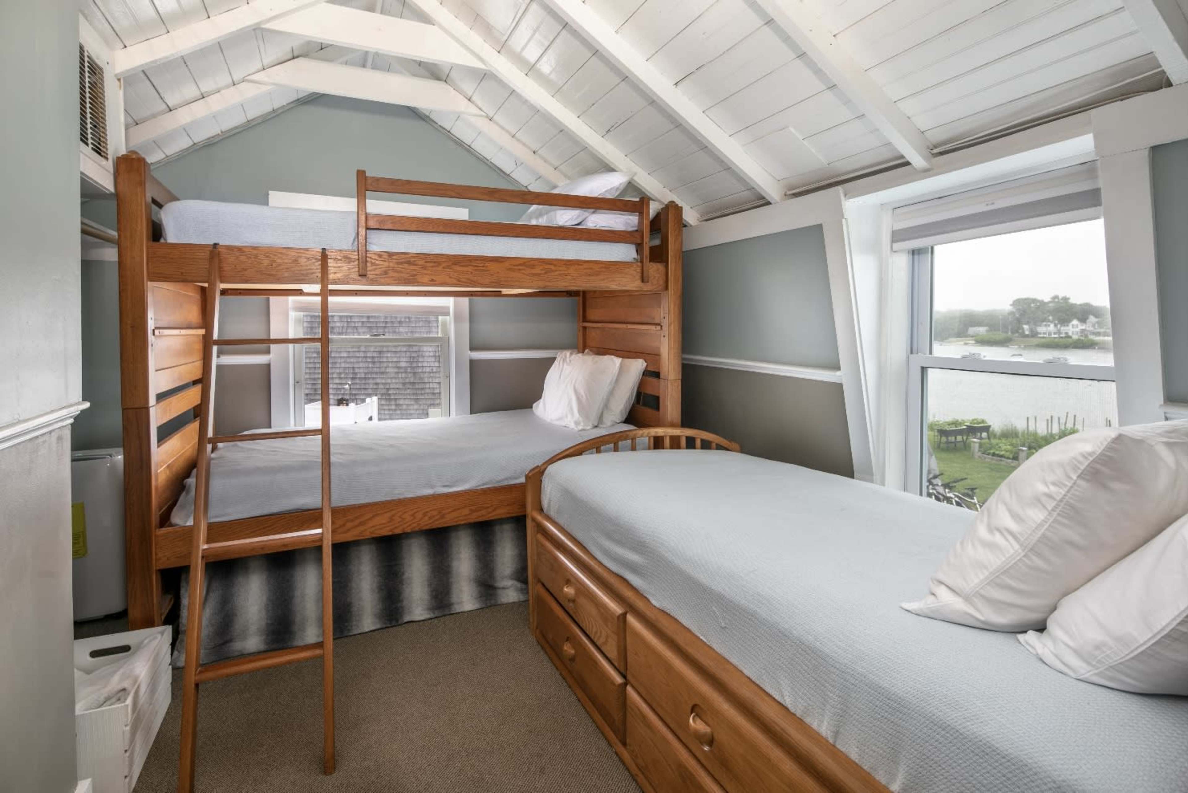 A small bedroom with a wooden bunk bed and a single bed, creating a cozy sleeping arrangement near a window with a view outside.