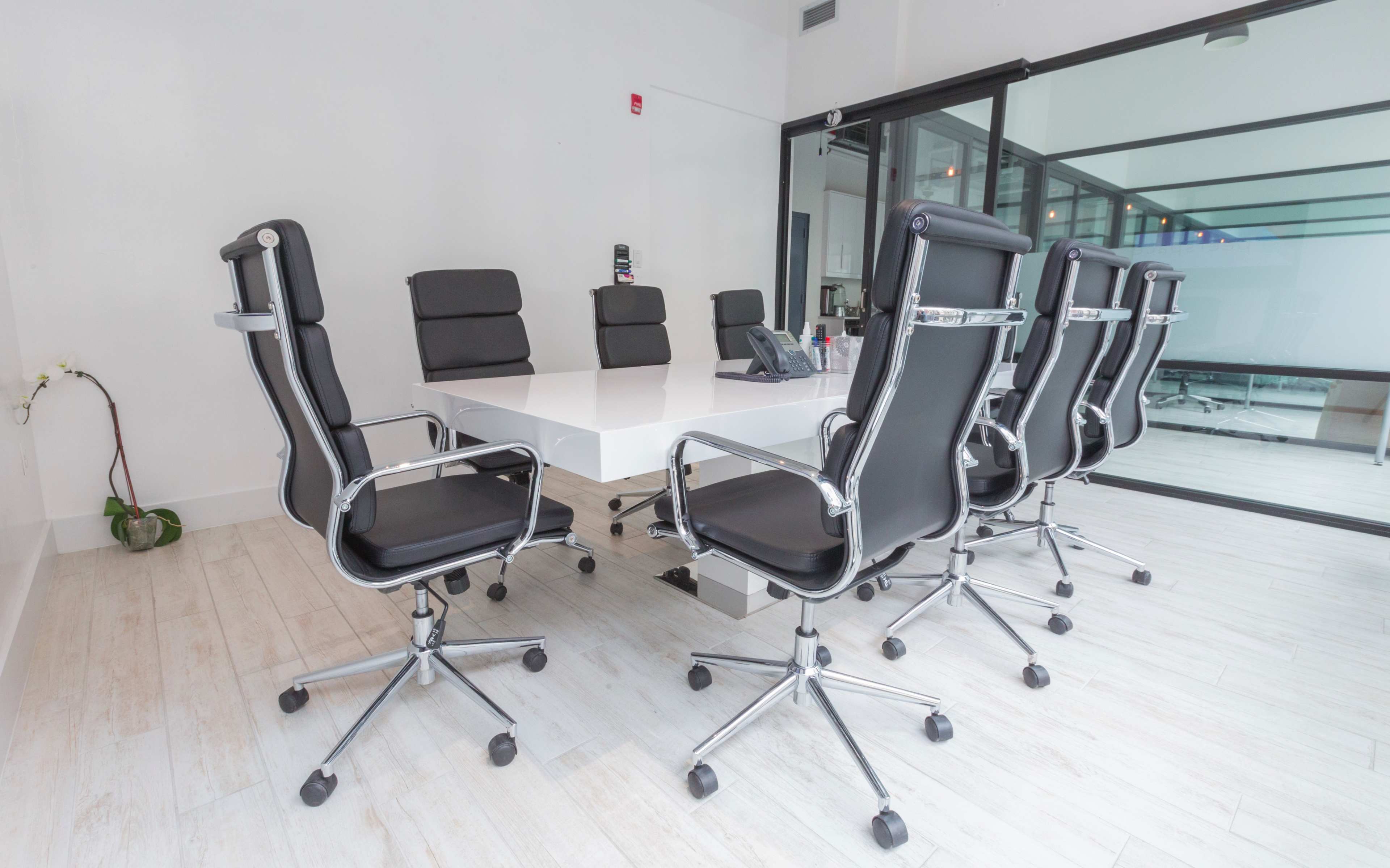 A modern conference room features a long white table surrounded by black ergonomic chairs and glass walls.