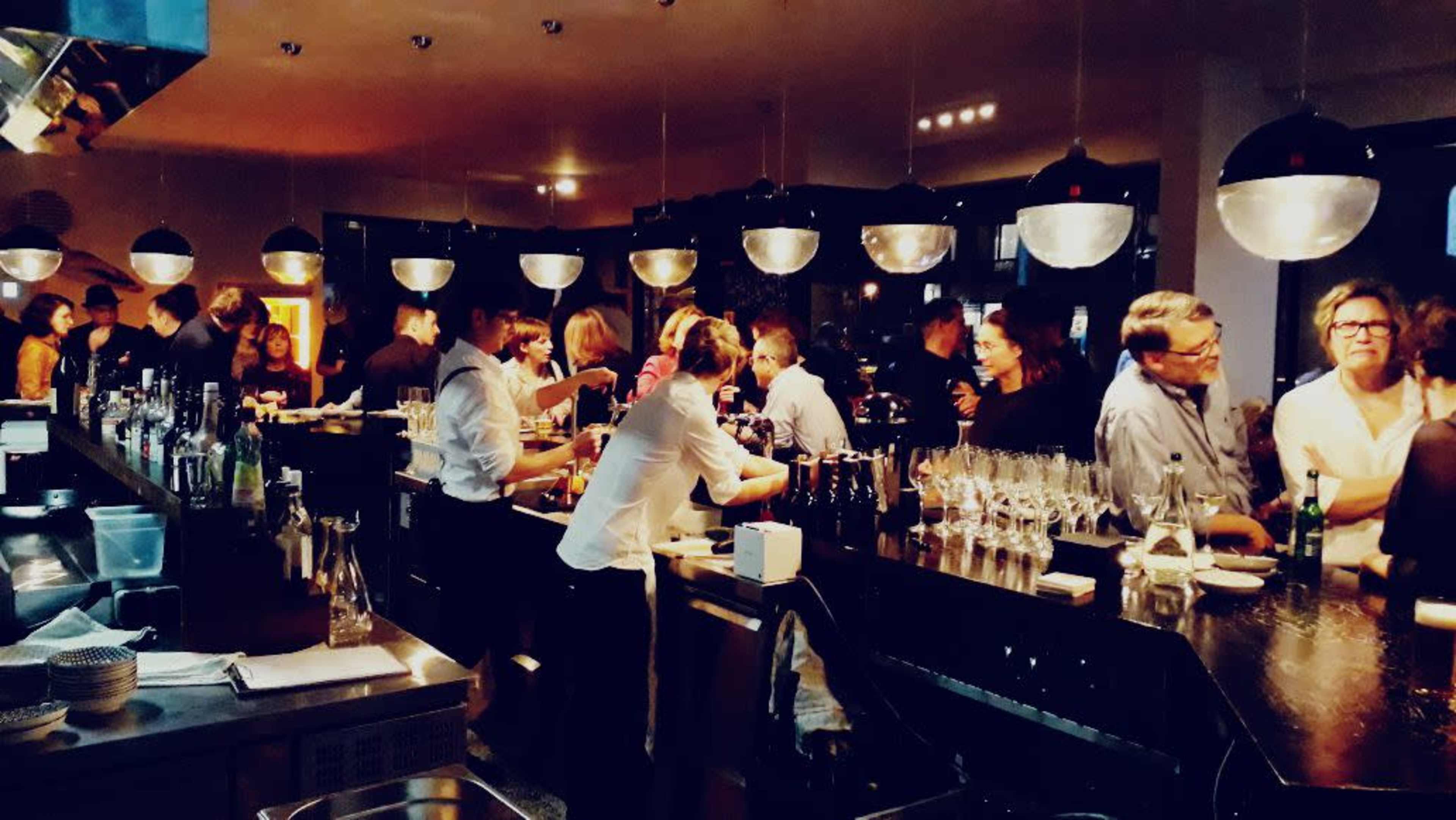 A busy bar scene filled with patrons socializing and bartenders preparing drinks under modern lighting.
