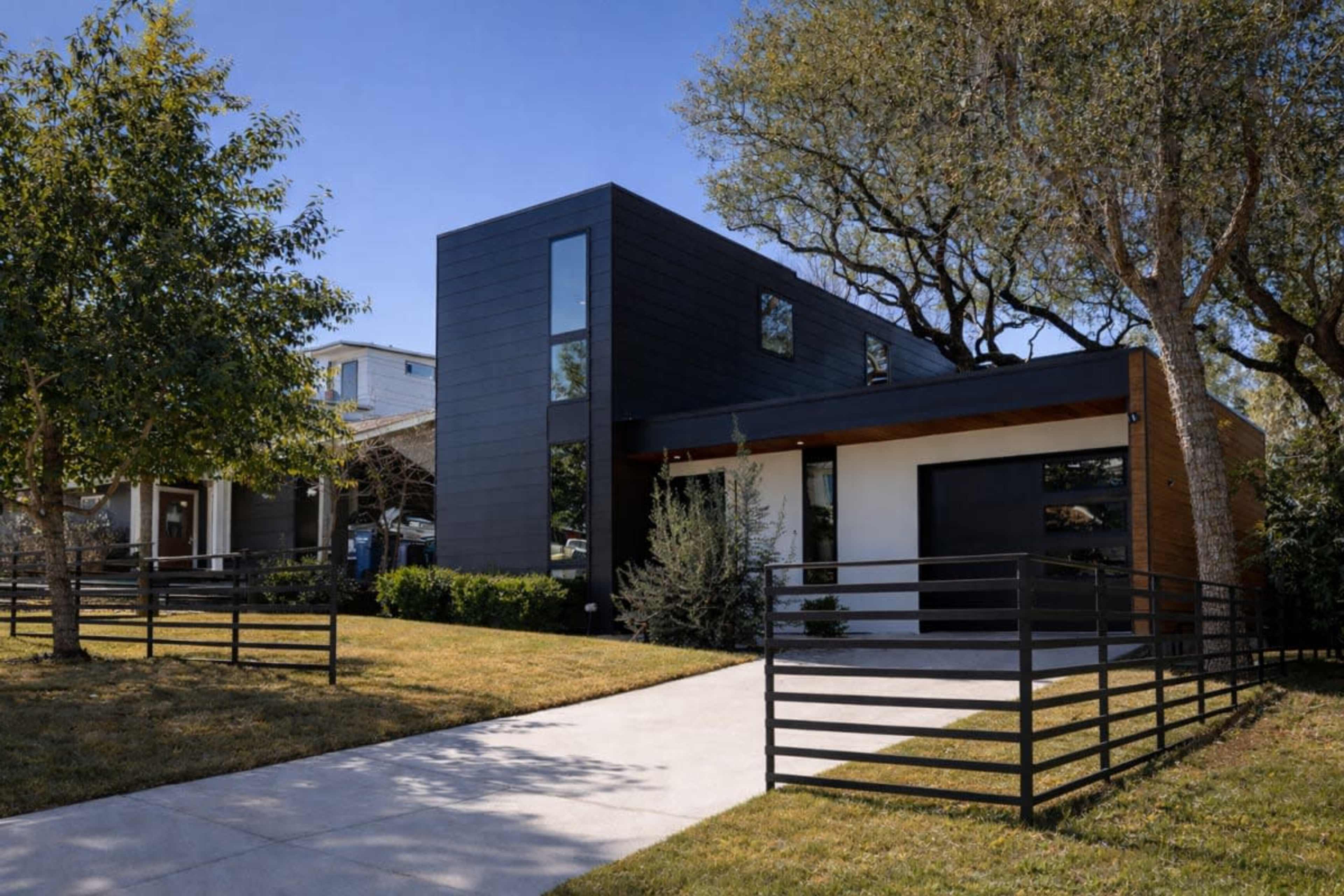 The image shows a modern two-story house with a black exterior and large windows, situated on a landscaped property with a concrete walkway and trees.