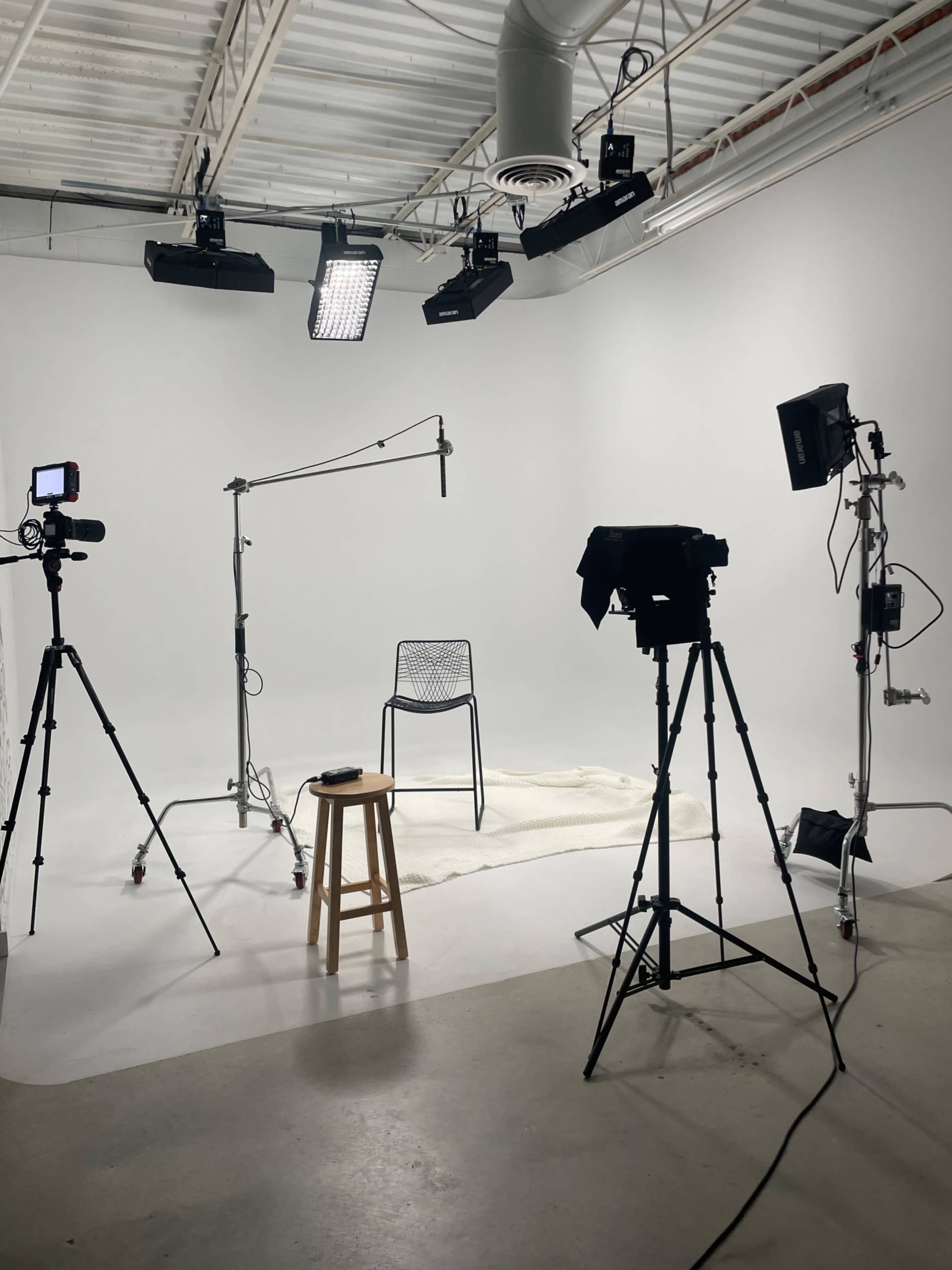 A minimalist filming studio features a single chair on a rug, surrounded by cameras and lighting equipment.