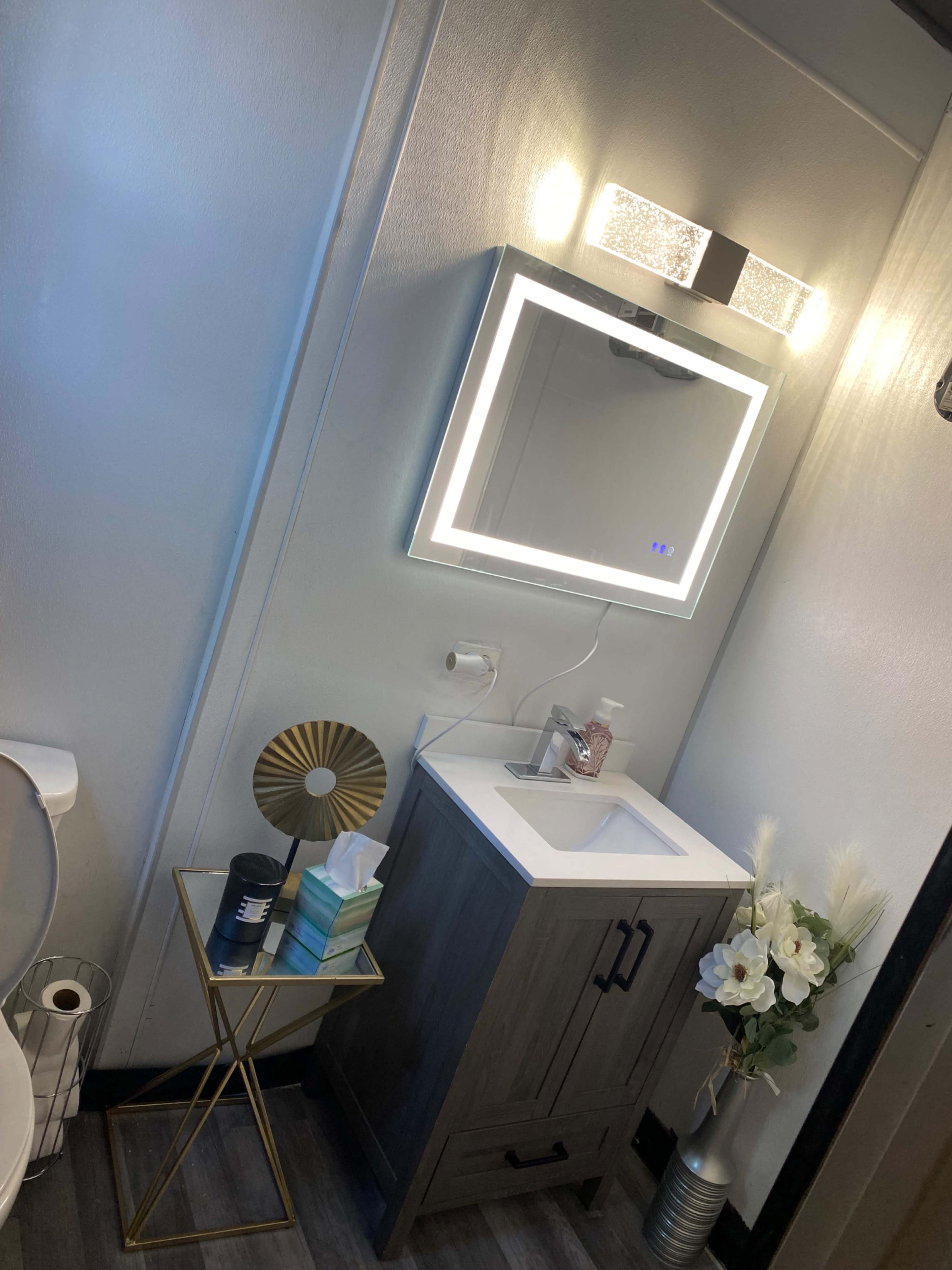 A modern bathroom with a backlit mirror above a wooden vanity, a decorative gold side table, and ceramic decorations.