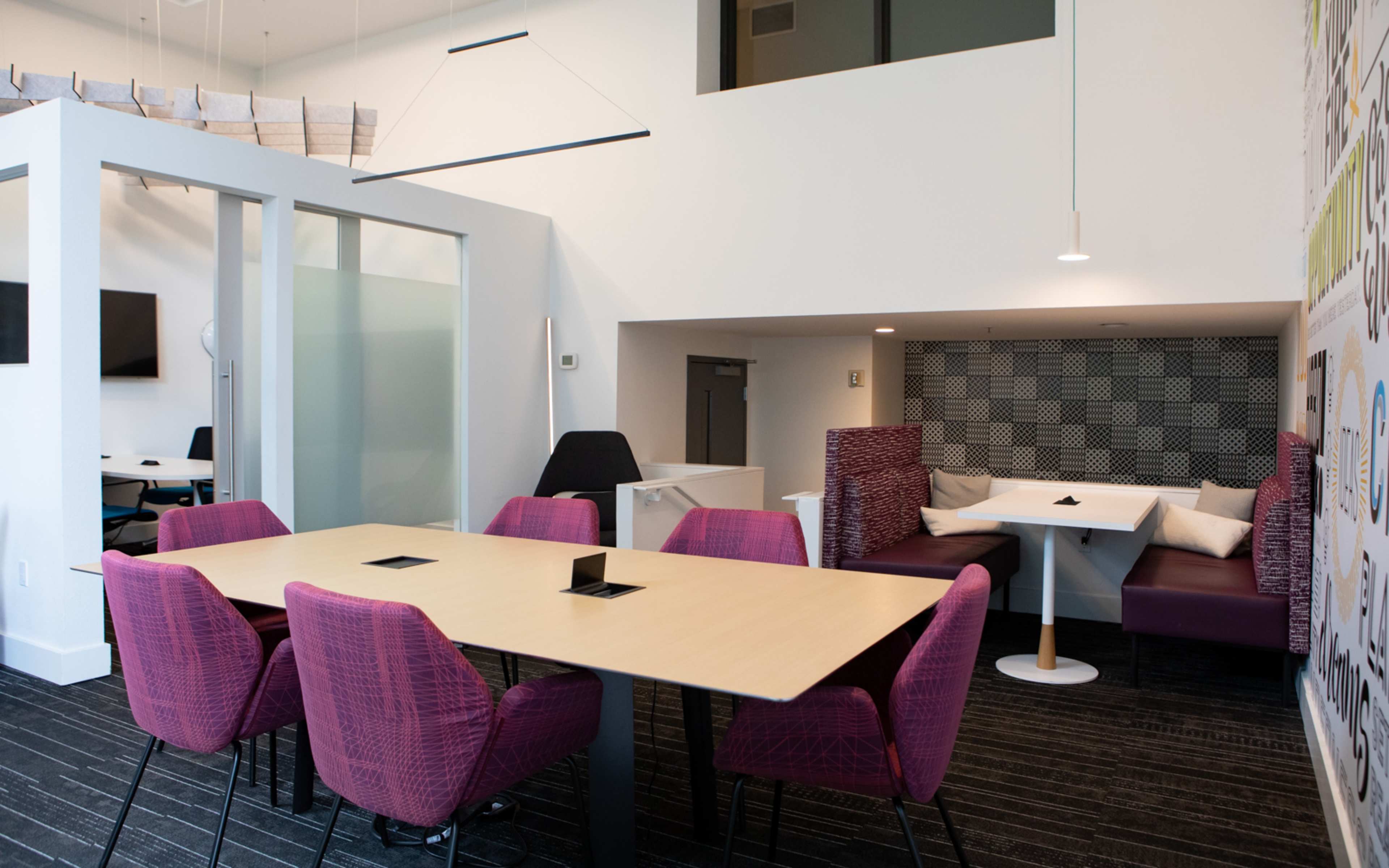 A modern office space featuring a conference-style table surrounded by pink chairs, with a booth area and a wall that includes a graphic design.