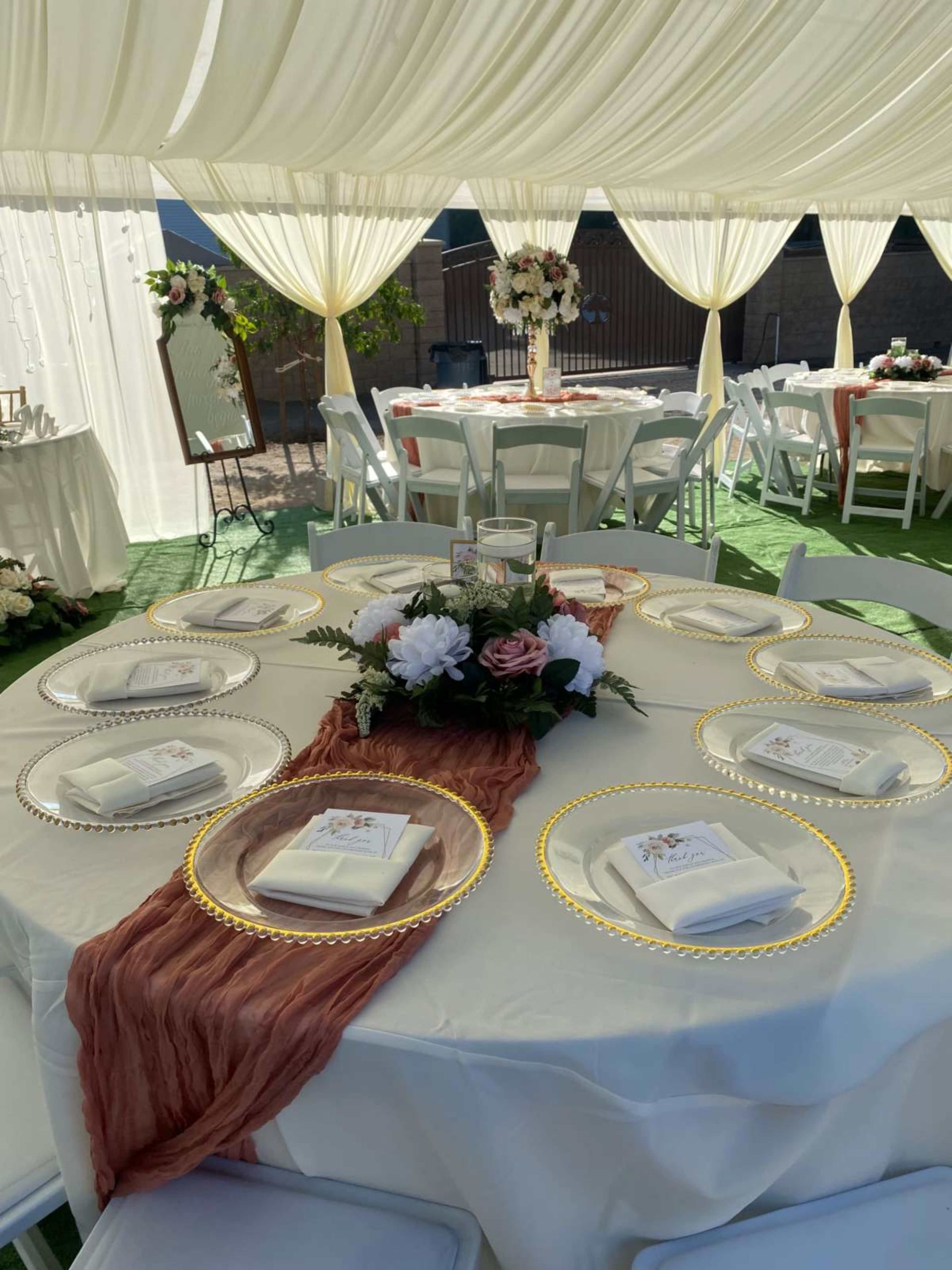 A decorated table is set with elegant plates and folded napkins under a canopy, surrounded by additional tables and floral arrangements.