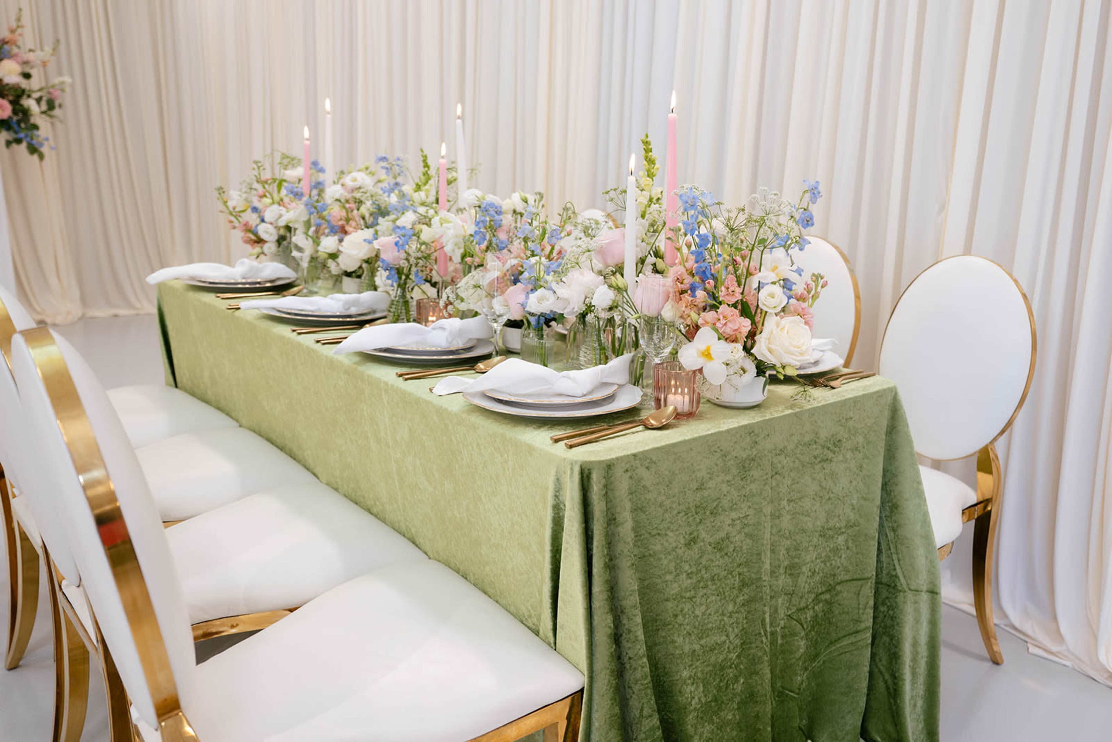 A long dining table is set with a green tablecloth, white dishware, and floral centerpieces, surrounded by gold-accented chairs.