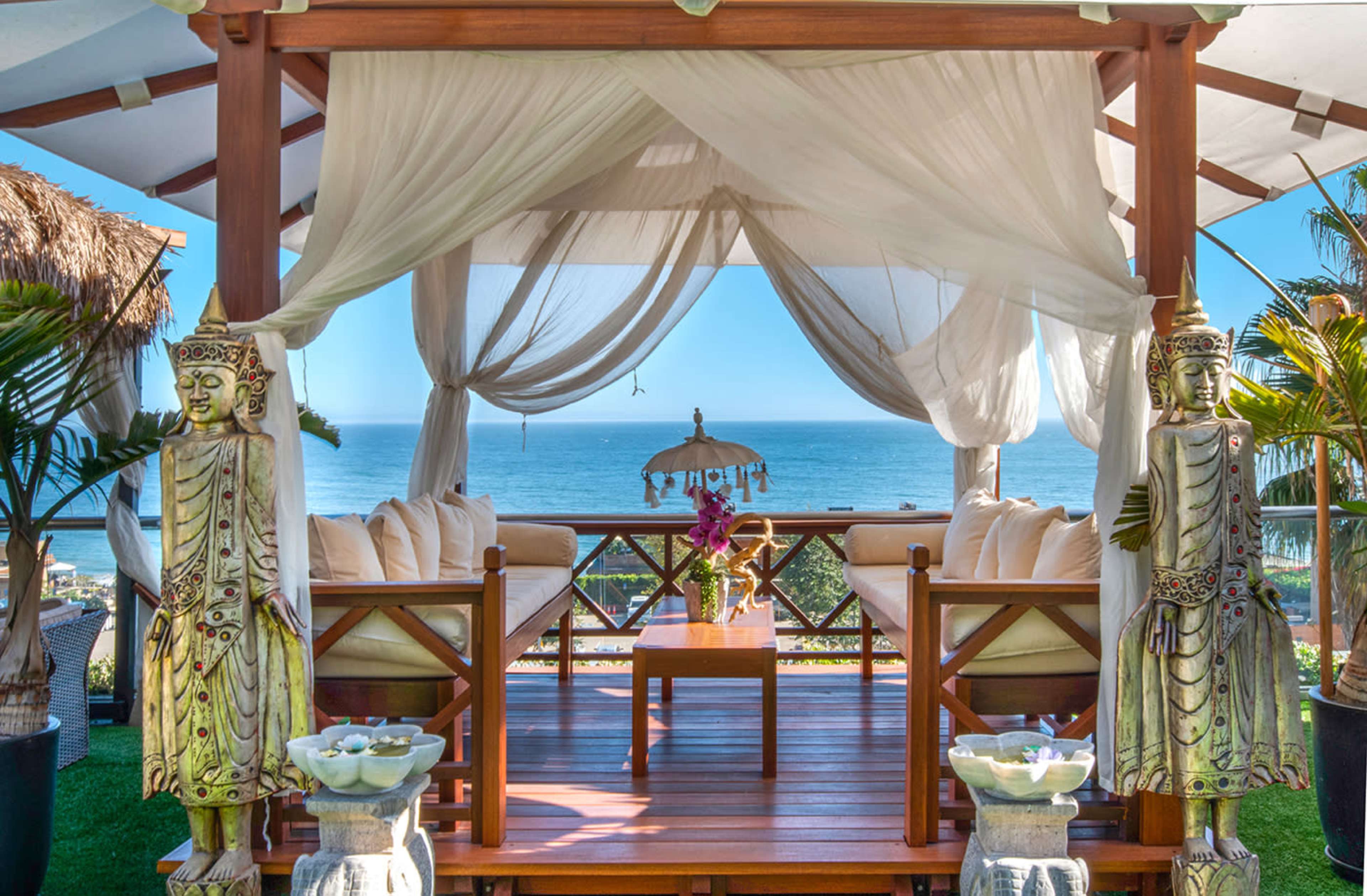 A wooden pavilion with draped fabric and a seated area overlooks the ocean, framed by decorative statues.