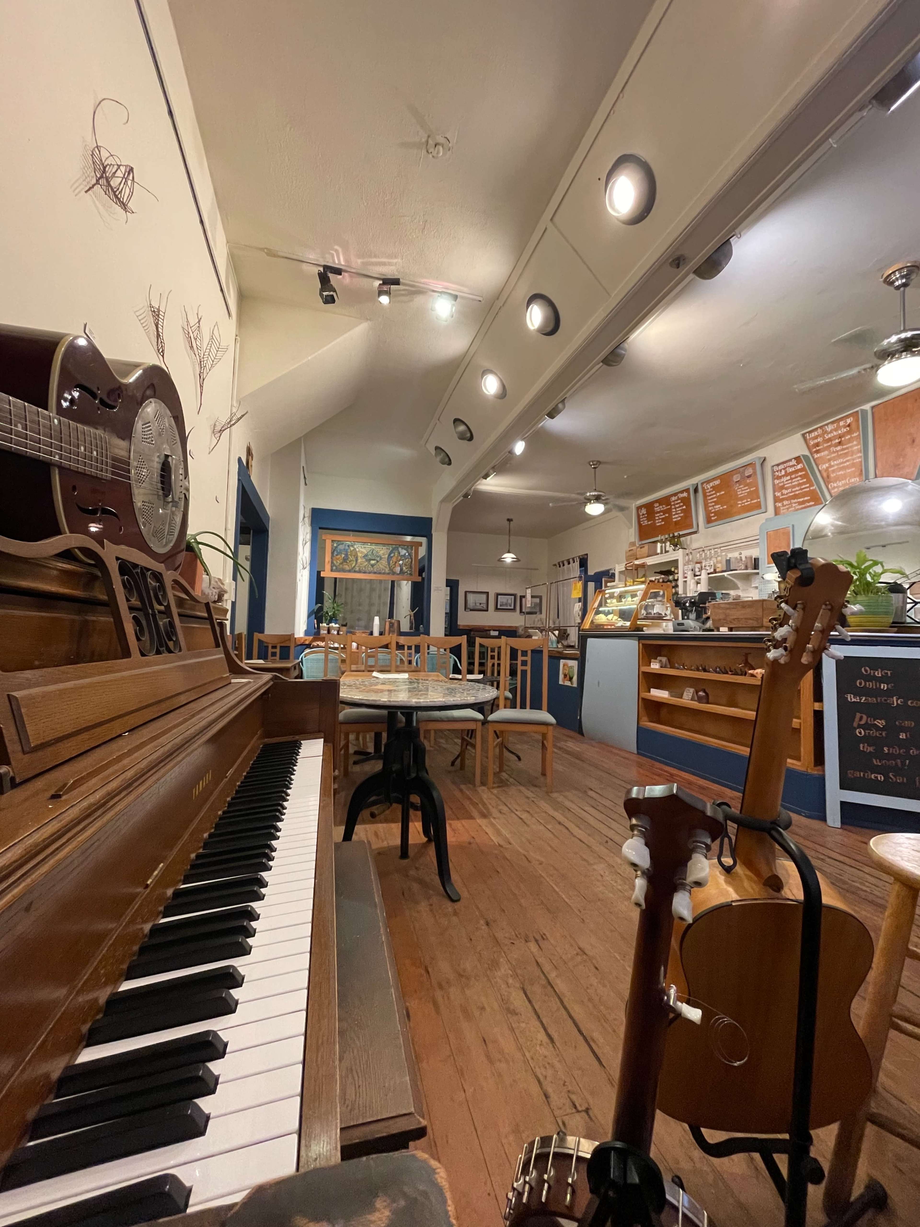 A cozy cafe interior featuring a wooden piano, tables, and guitars, with a counter displaying a menu on the back wall.