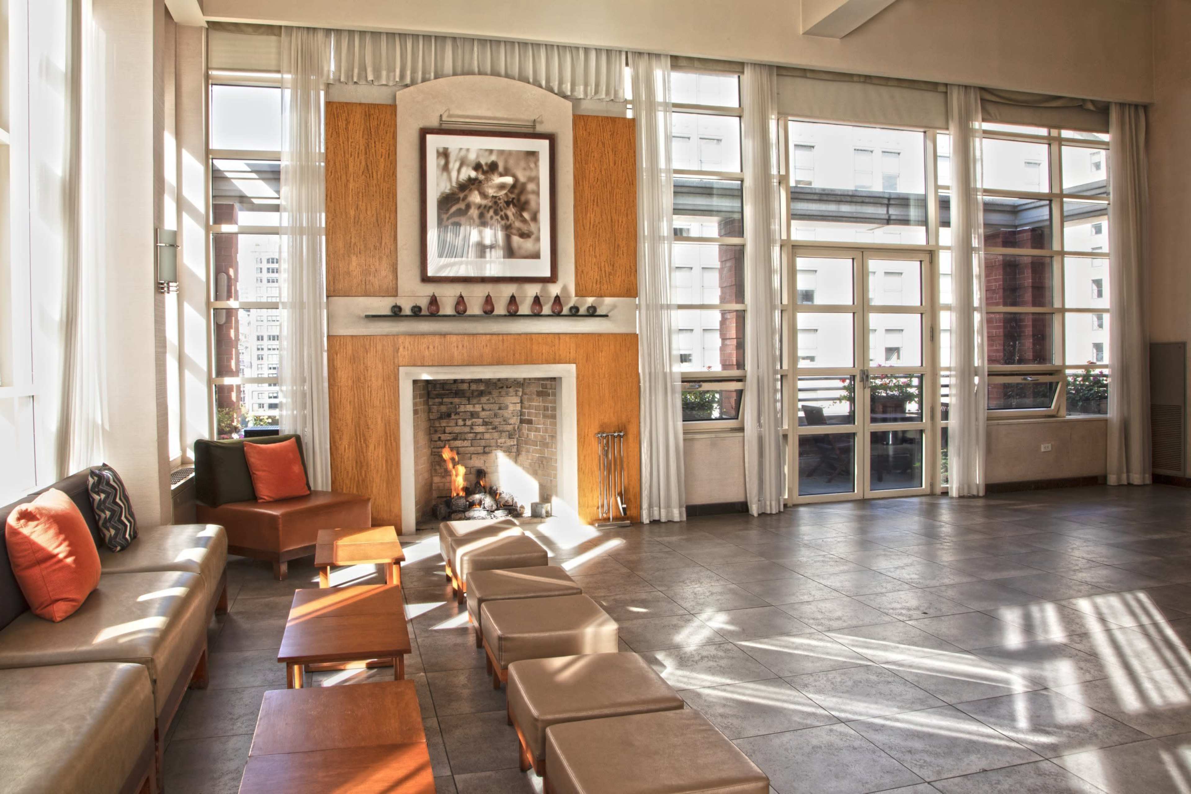 The image shows a modern lounge area with a fireplace, large windows allowing natural light, and seating arranged around small tables.