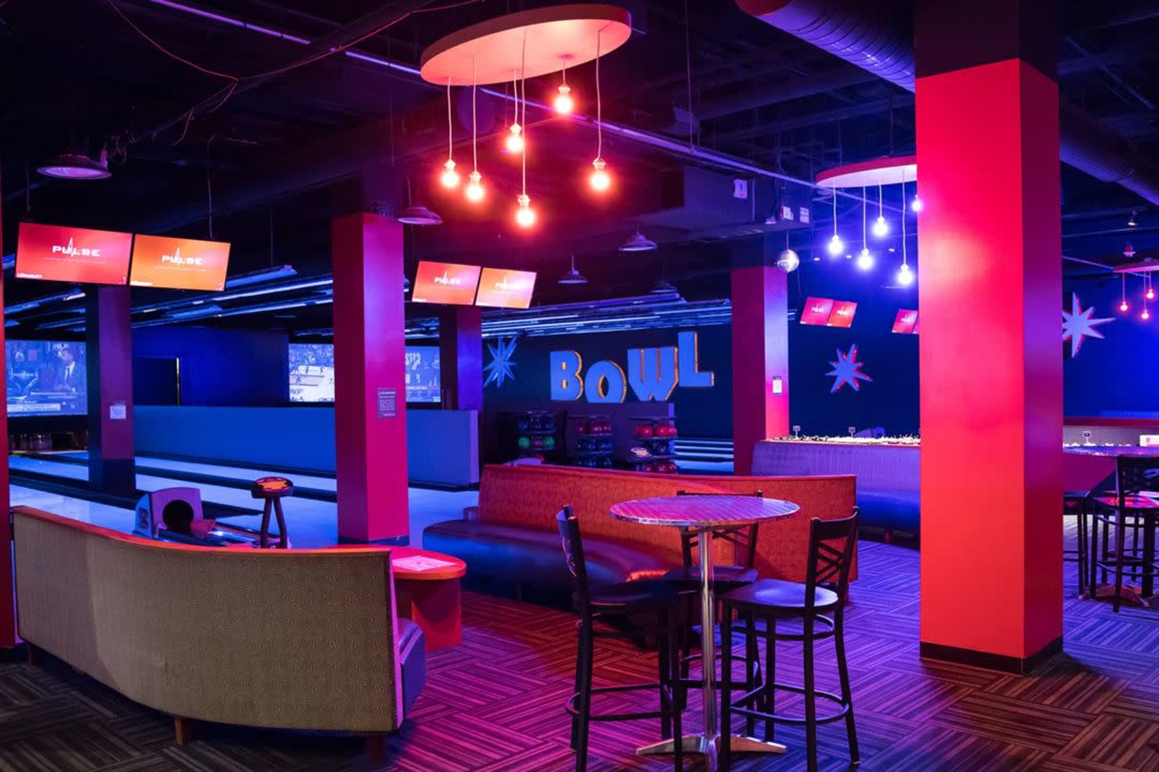 The image shows a bowling alley interior with vibrant lighting, including colorful pendant lights, seating areas, and bowling lanes in the background.