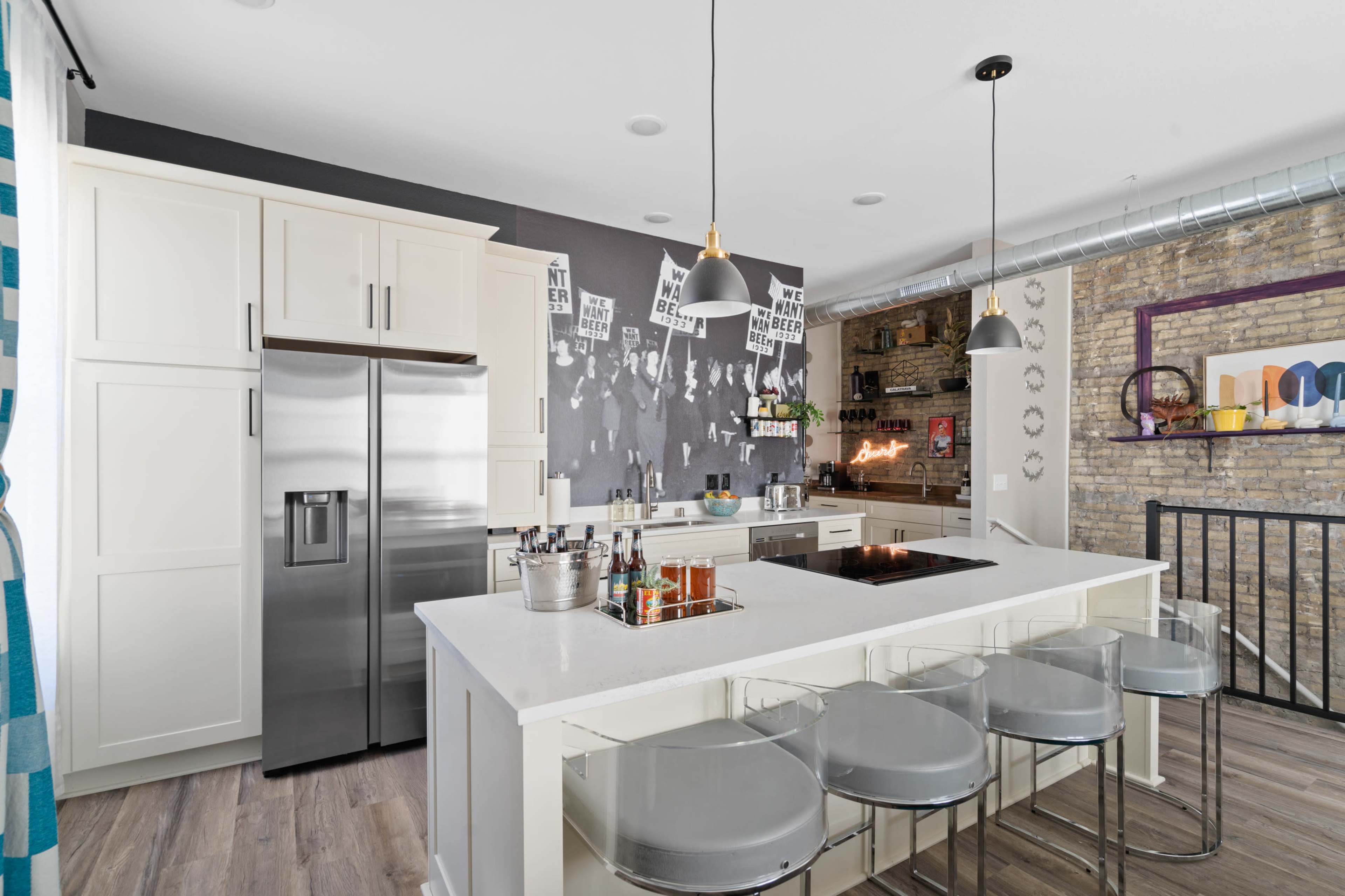 A modern kitchen features a gray accent wall with posters, stainless steel appliances, a large island with bar stools, and wood flooring.