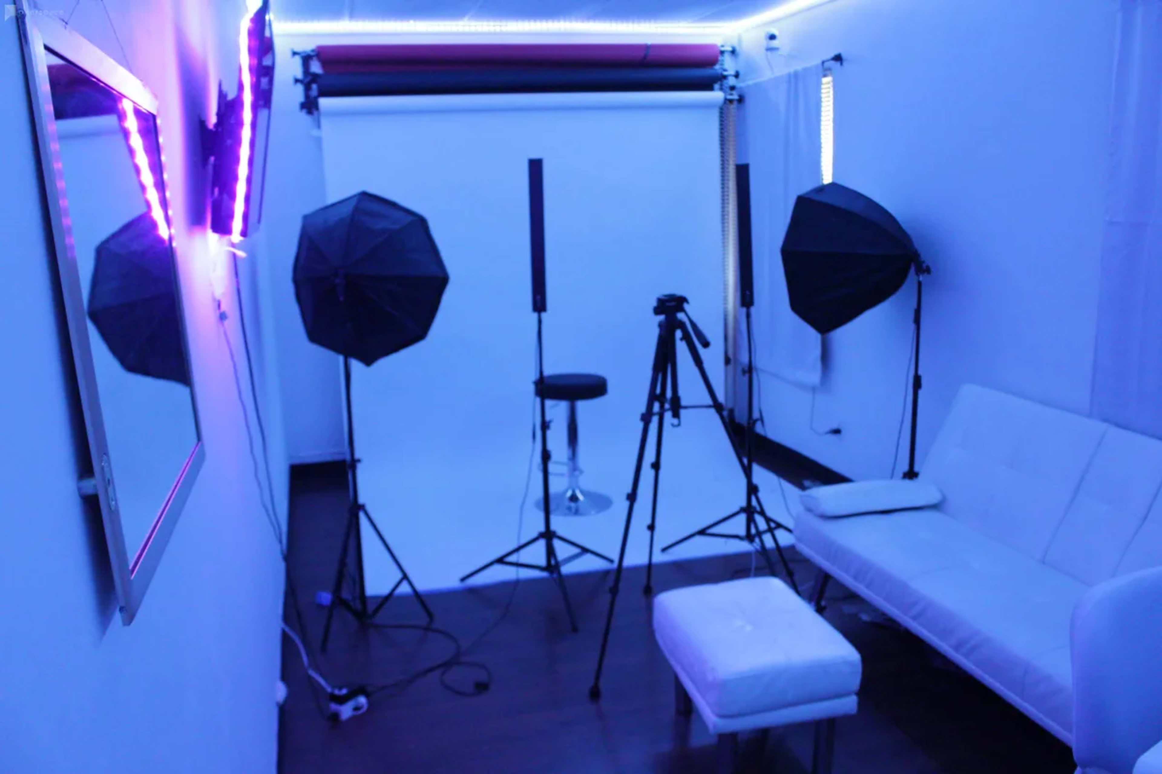 A photography studio featuring a white backdrop, two softbox lights, a tripod, and modern furniture in blue lighting.
