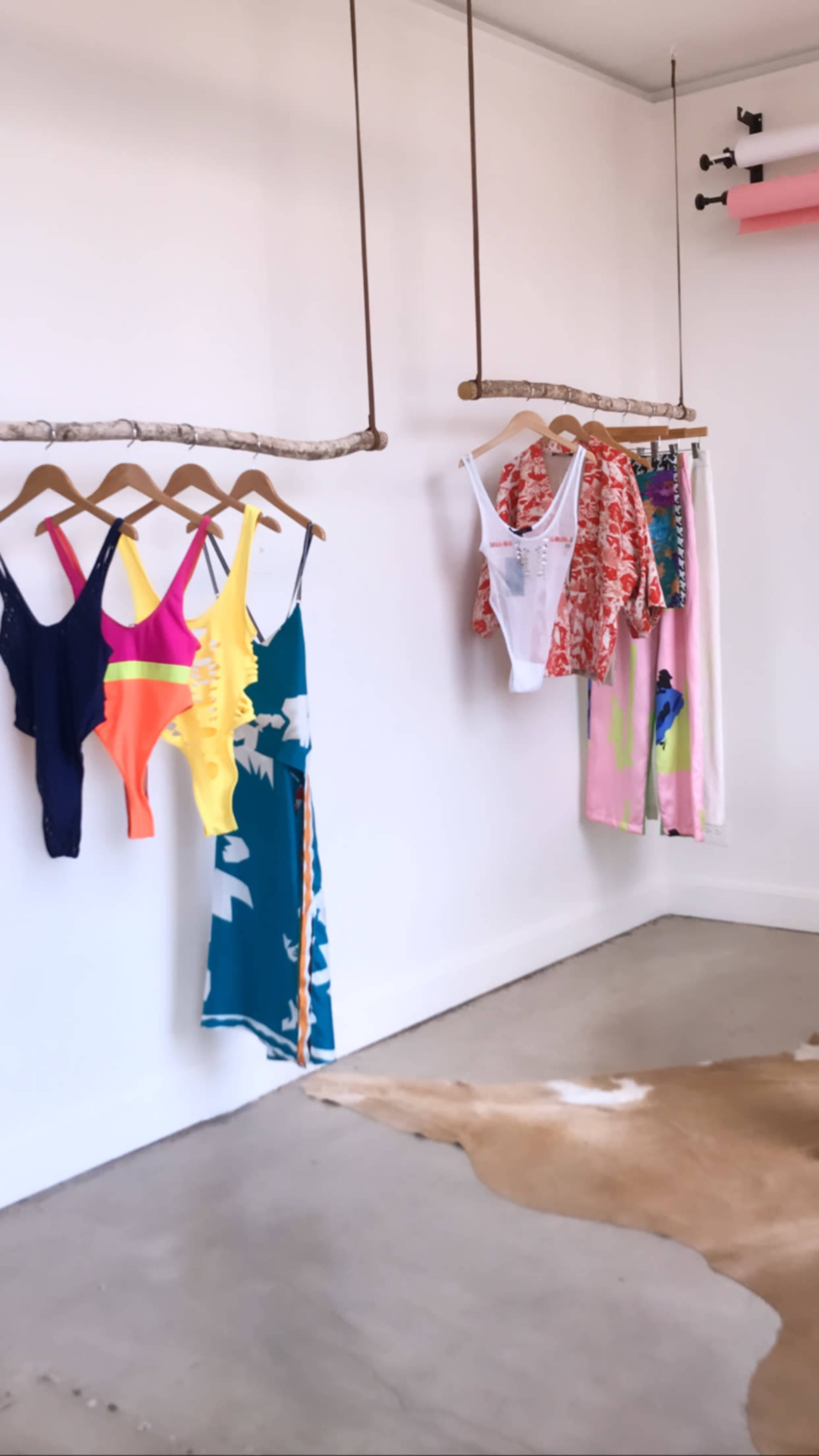 A clothing display with swimwear hanging on wooden rods against a white wall, complemented by a cowhide rug on the floor.