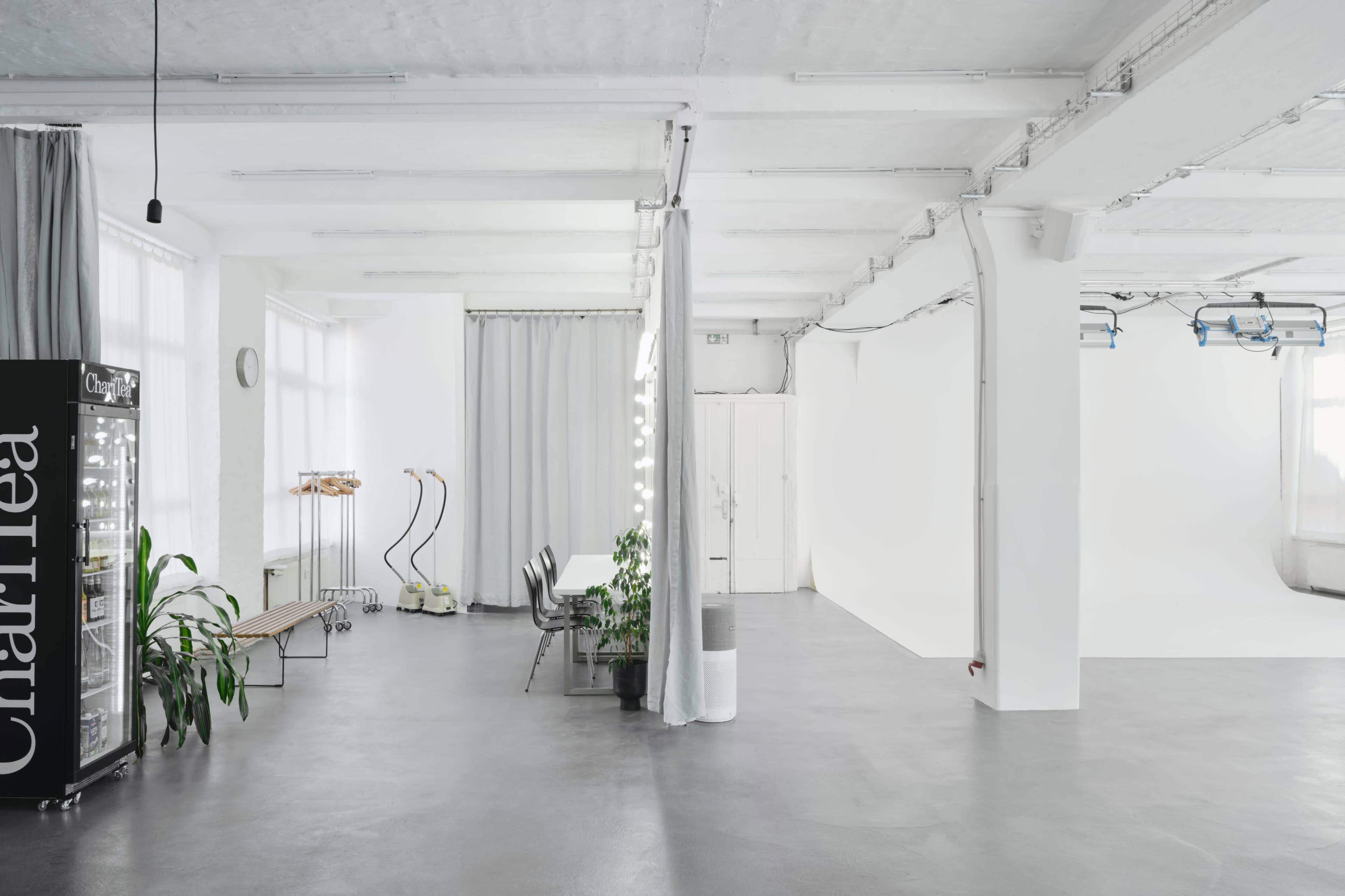 The image shows a spacious, minimalistic studio with white walls, concrete floors, and several curtains, alongside a clothing rack and potted plants.
