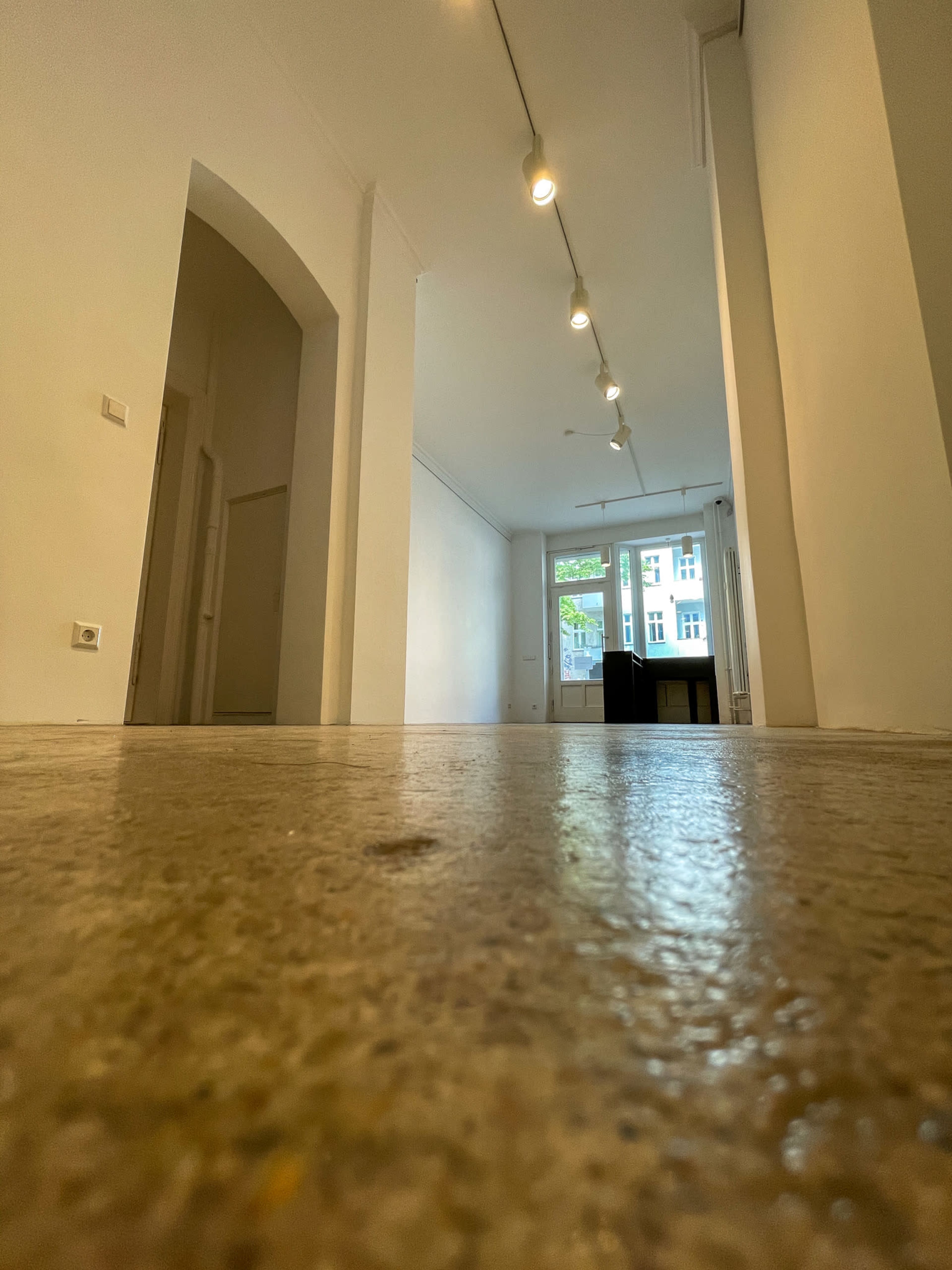 The image shows a spacious corridor leading to a bright room illuminated by overhead lights, with a polished concrete floor.