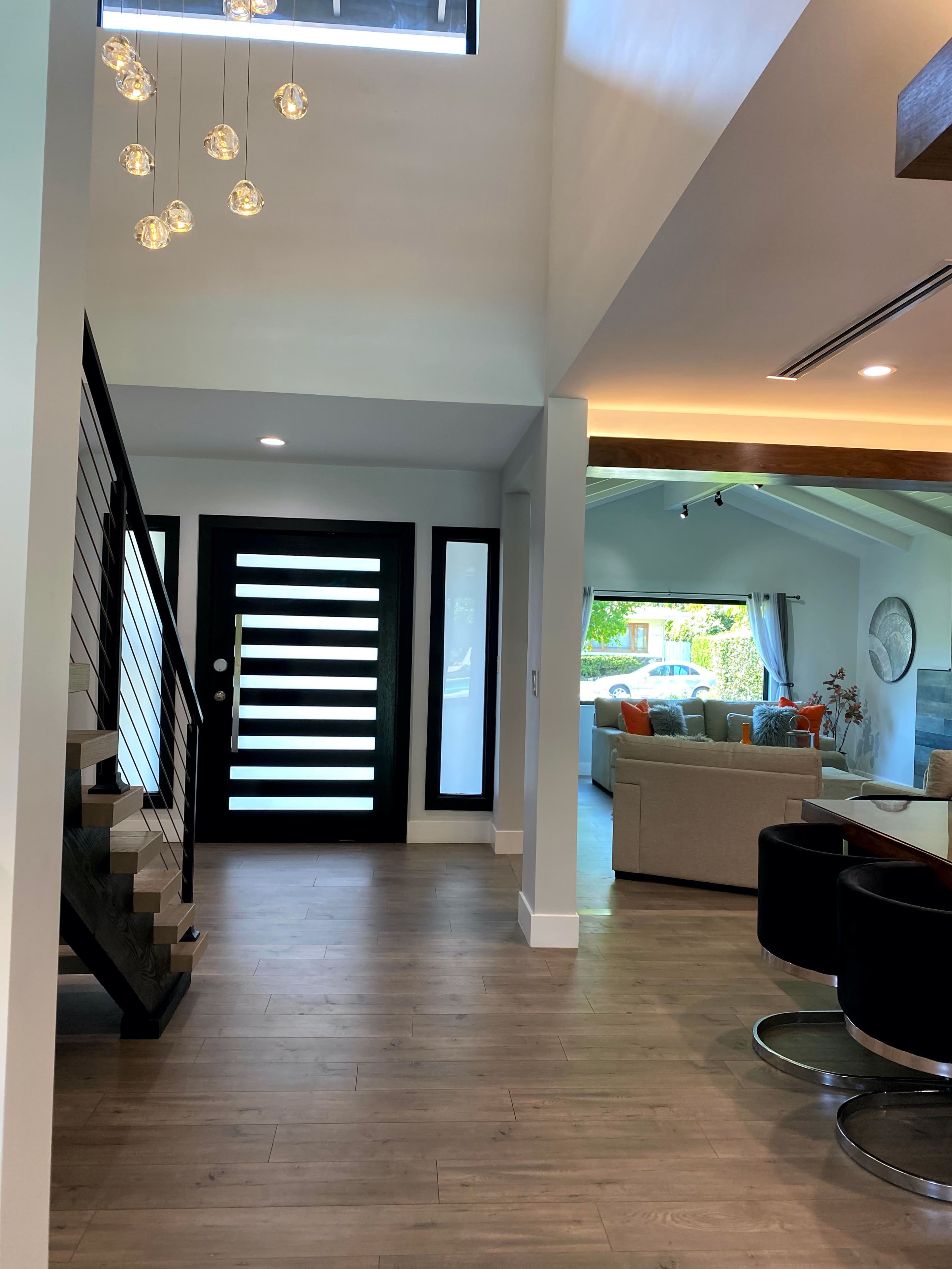A modern entryway with a staircase on the left, a large black front door with horizontal glass panels, and a view into a living area with a sofa visible in the background.