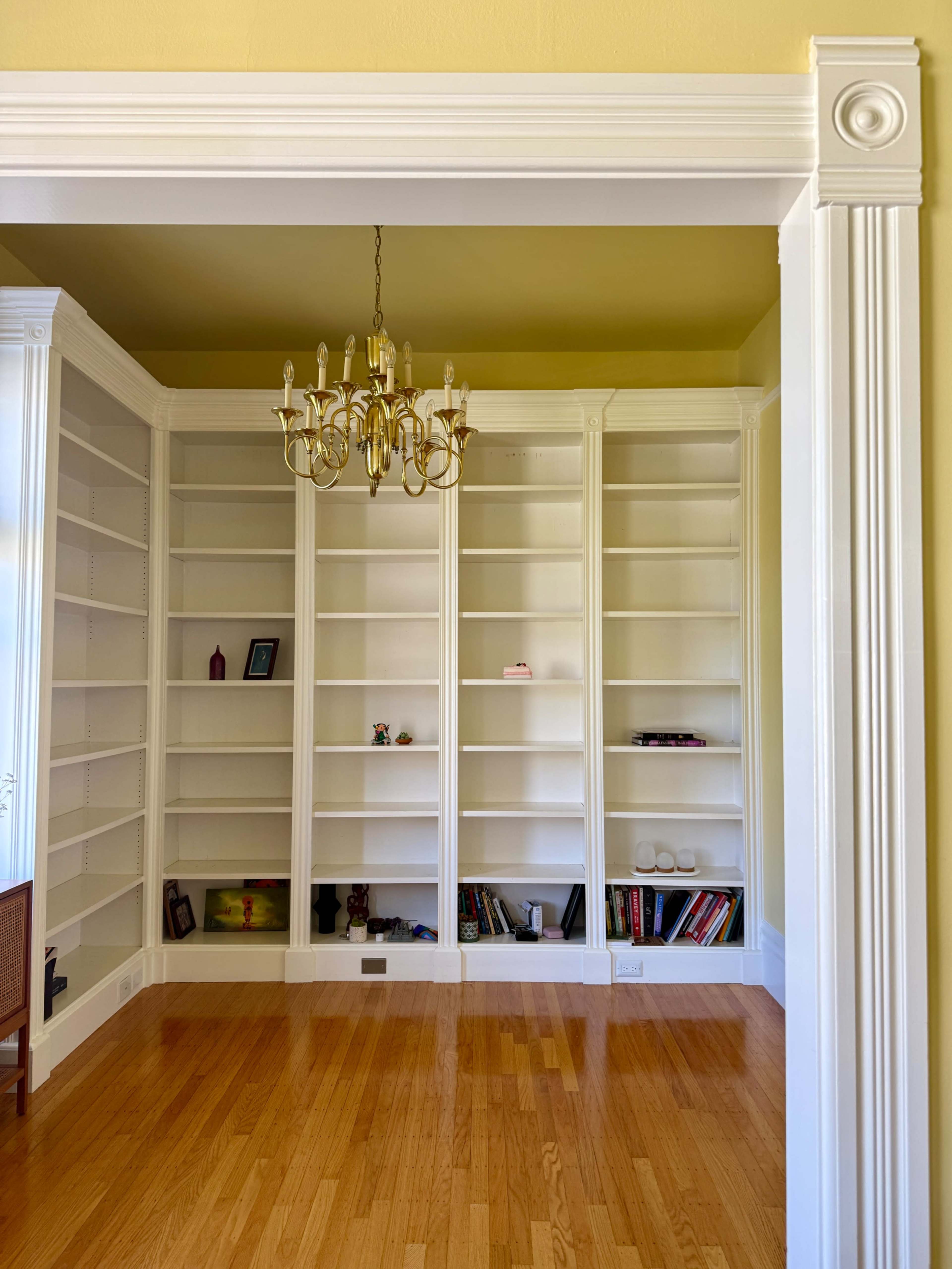 The image shows an empty room with built-in white bookshelves and a chandelier, featuring a light yellow wall and polished wooden flooring.