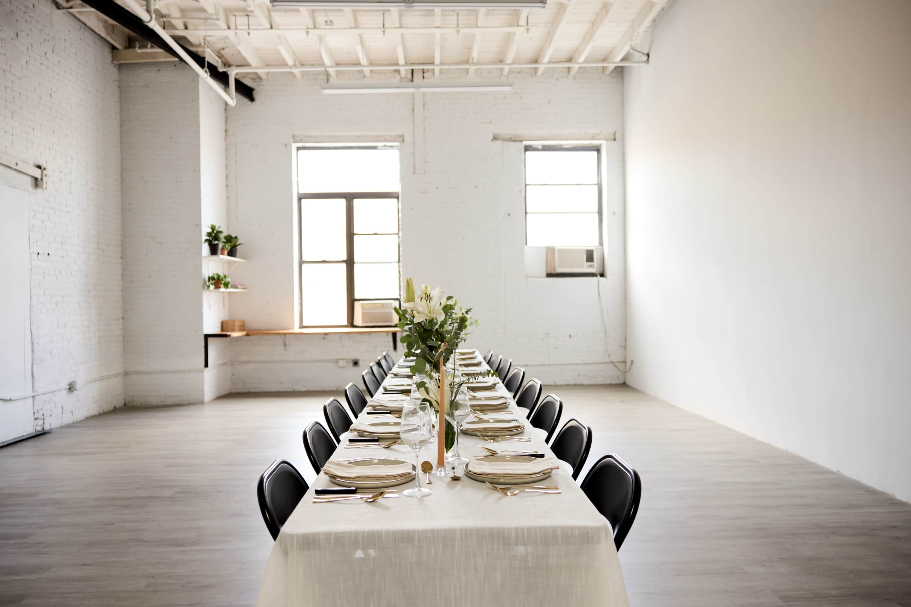 A long table is set with dinnerware and a decorative centerpiece, positioned in a spacious, well-lit room with large windows and minimal decor.