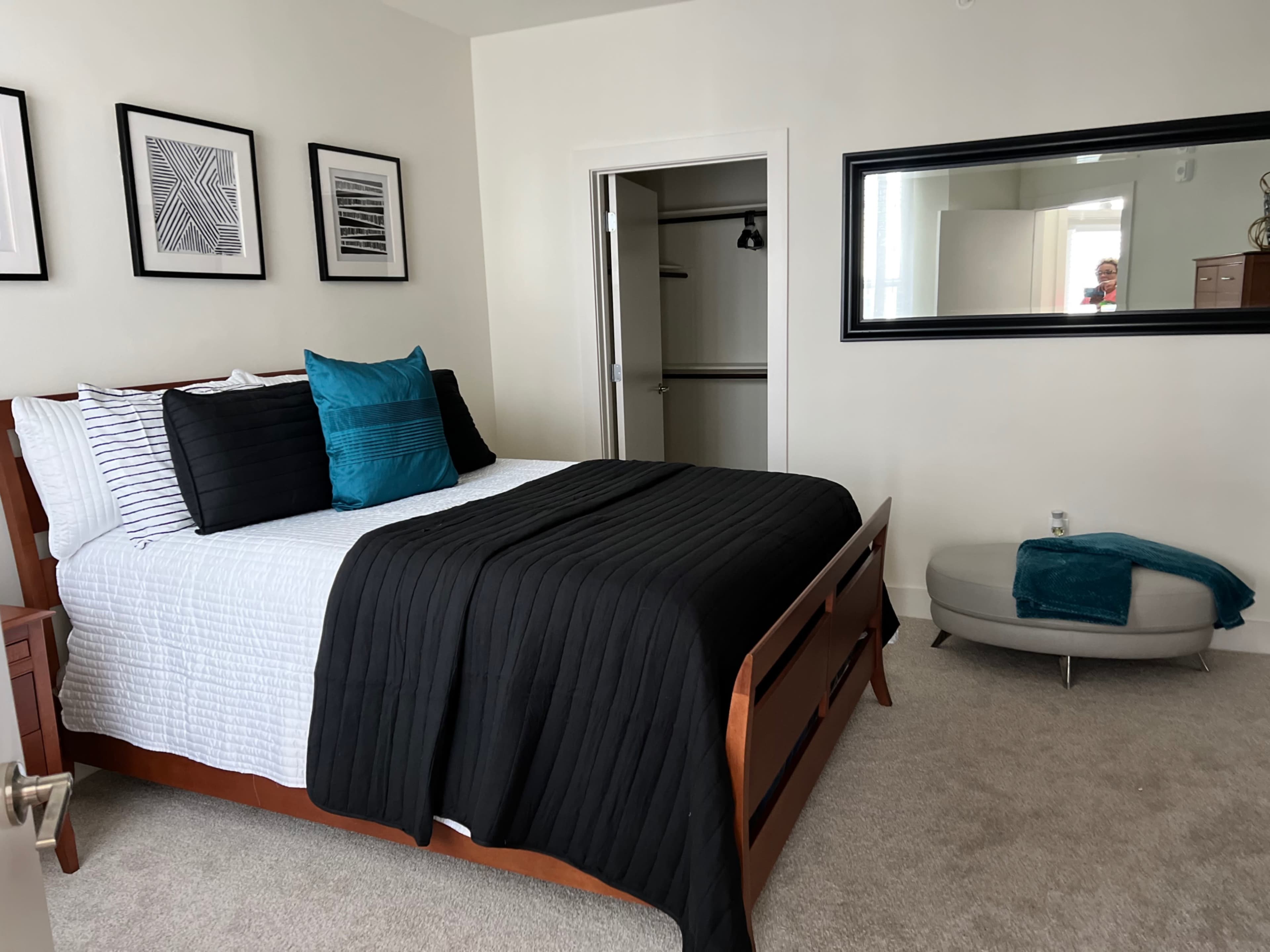 A neatly arranged bedroom features a large bed with black and white bedding, framed artwork on the walls, and a mirror above a small seating area.
