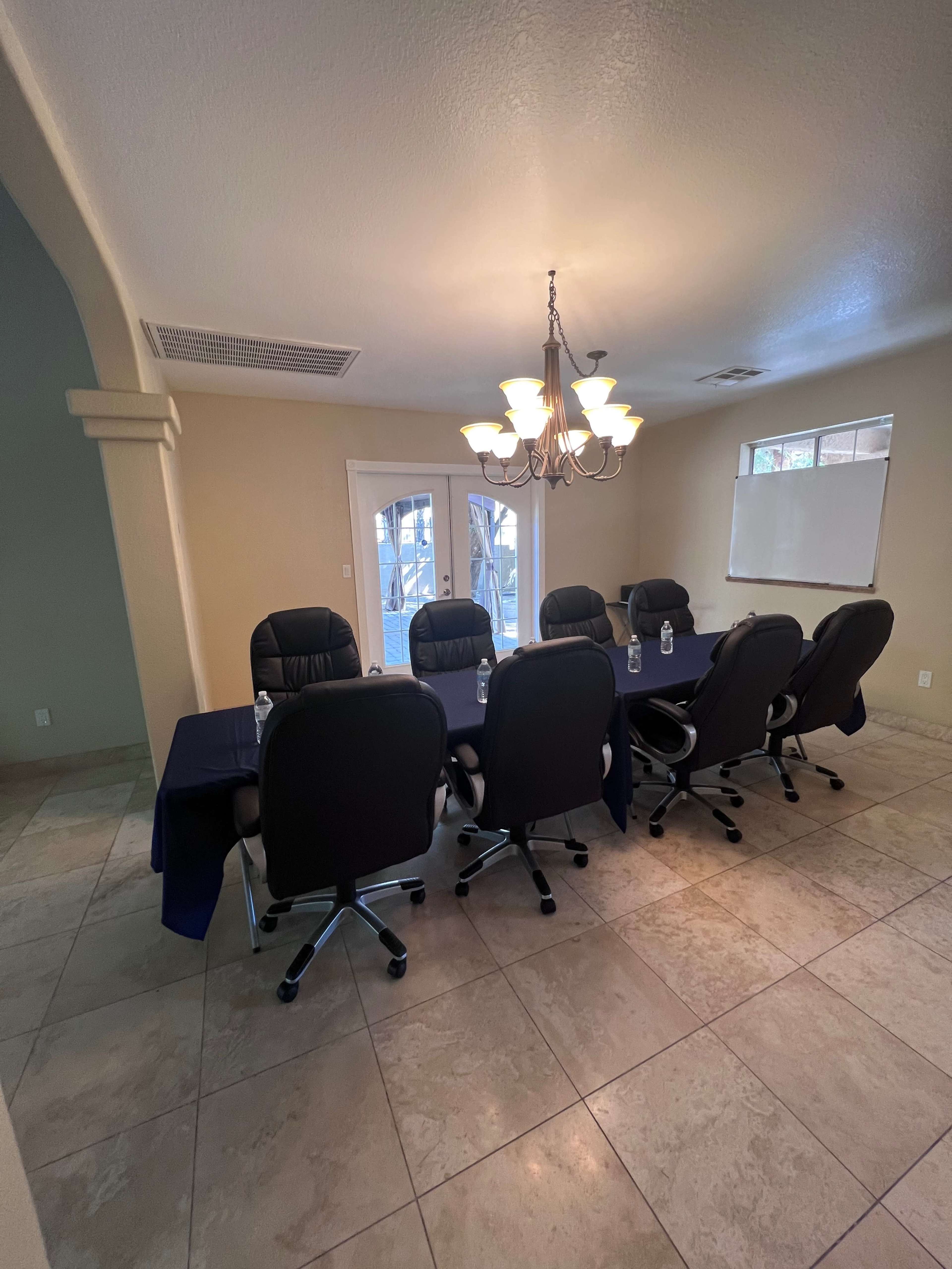 A conference room features a long table surrounded by six black office chairs, with a chandelier overhead and large windows providing natural light.