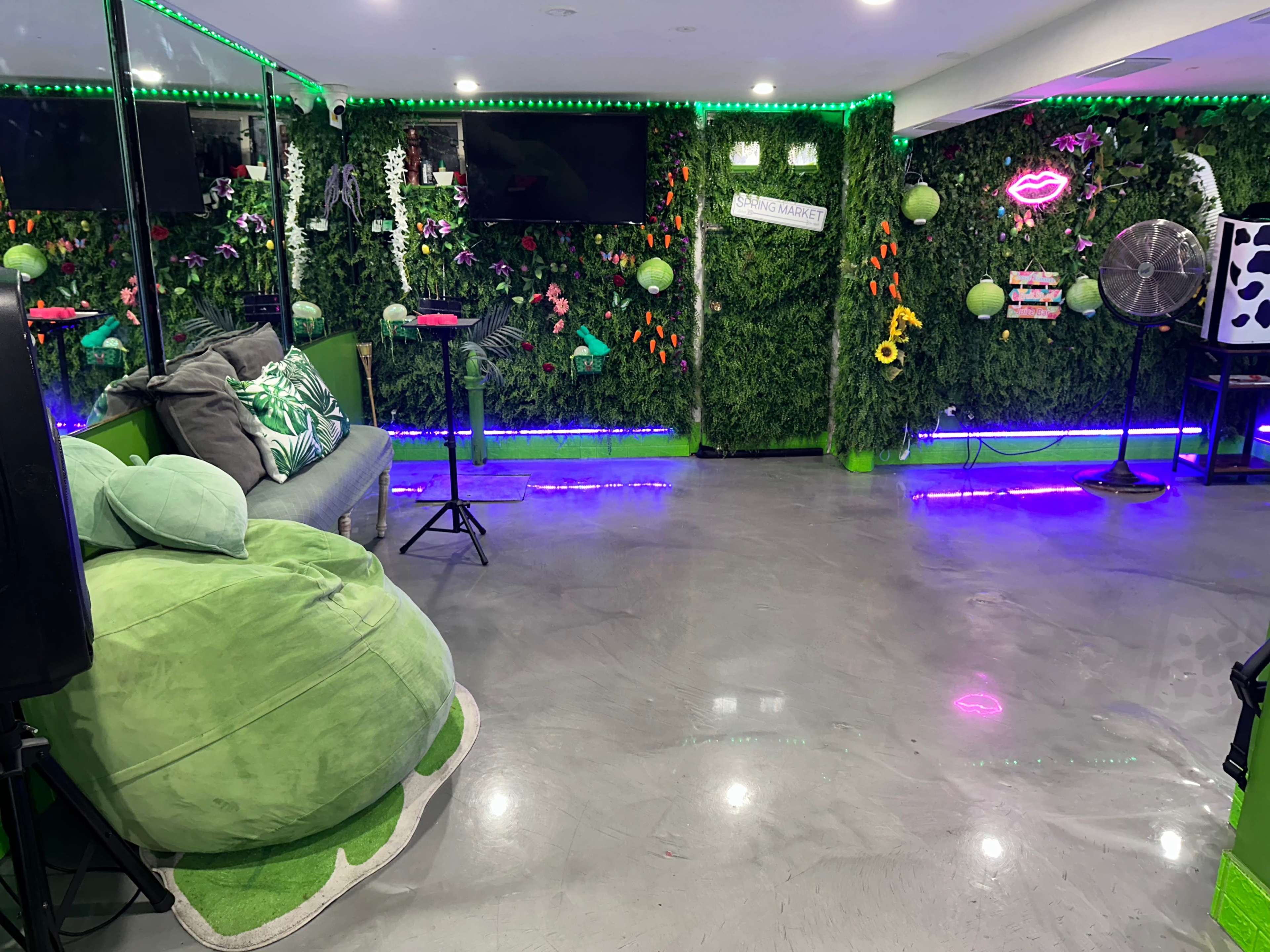 The room features green foliage walls, neon lighting, a couch, a bean bag chair, and a fan.