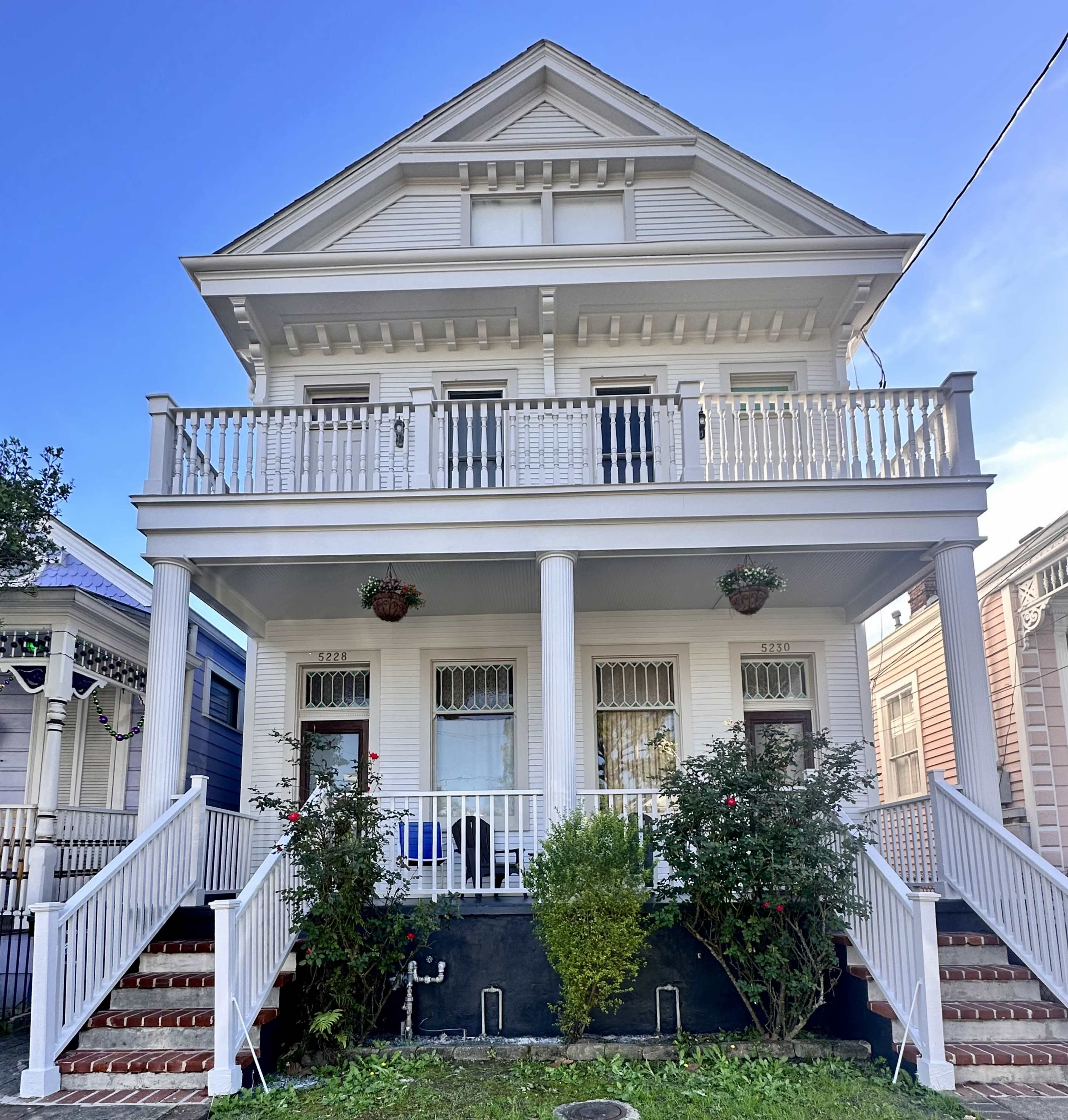 A two-story, white wooden house with a prominent balcony, two sets of stairs leading to the entrance, and flower pots hanging from the railing.