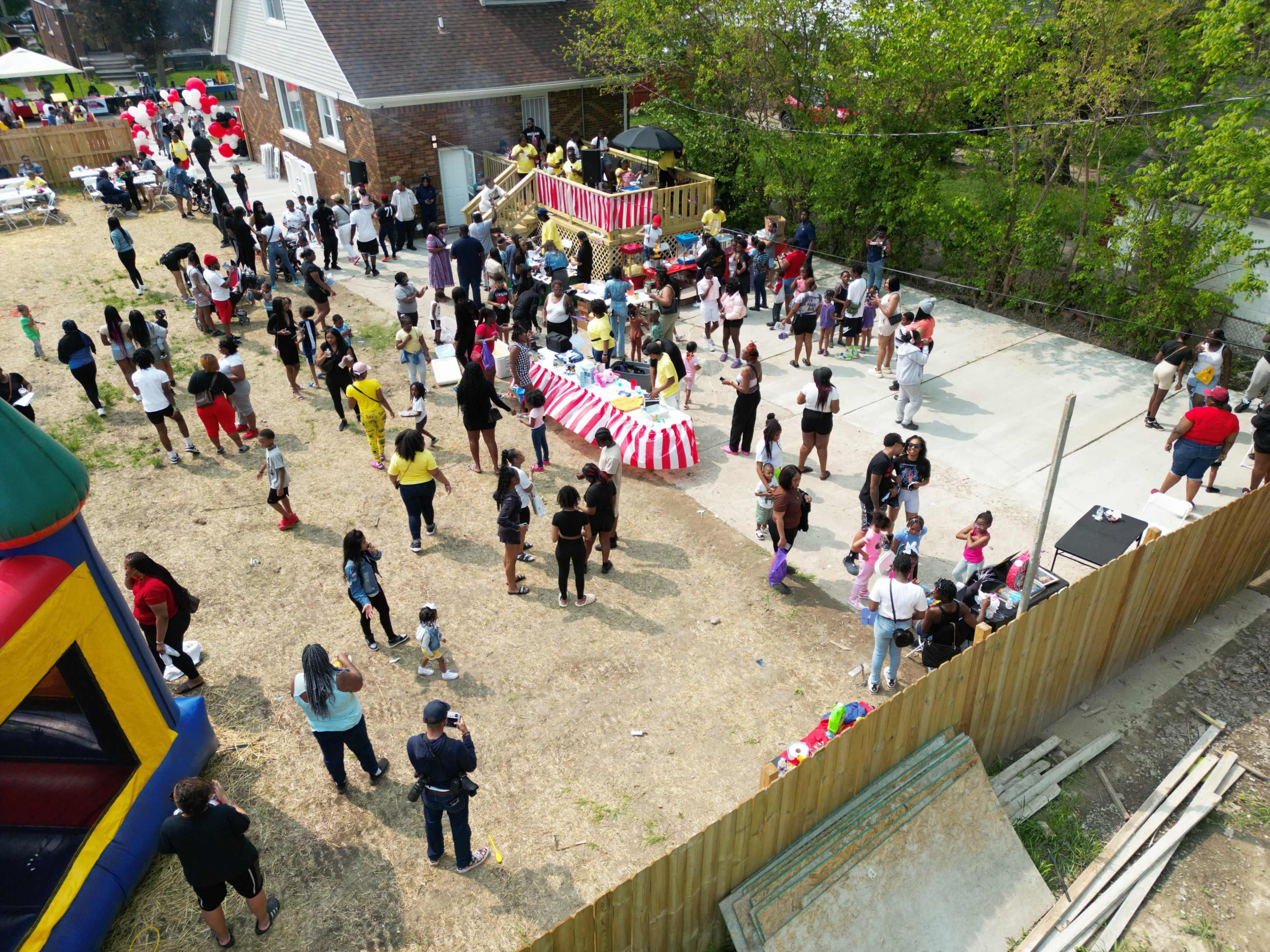 A large group of people gathers in a backyard with a bounce house, tables with food, and festive decorations.