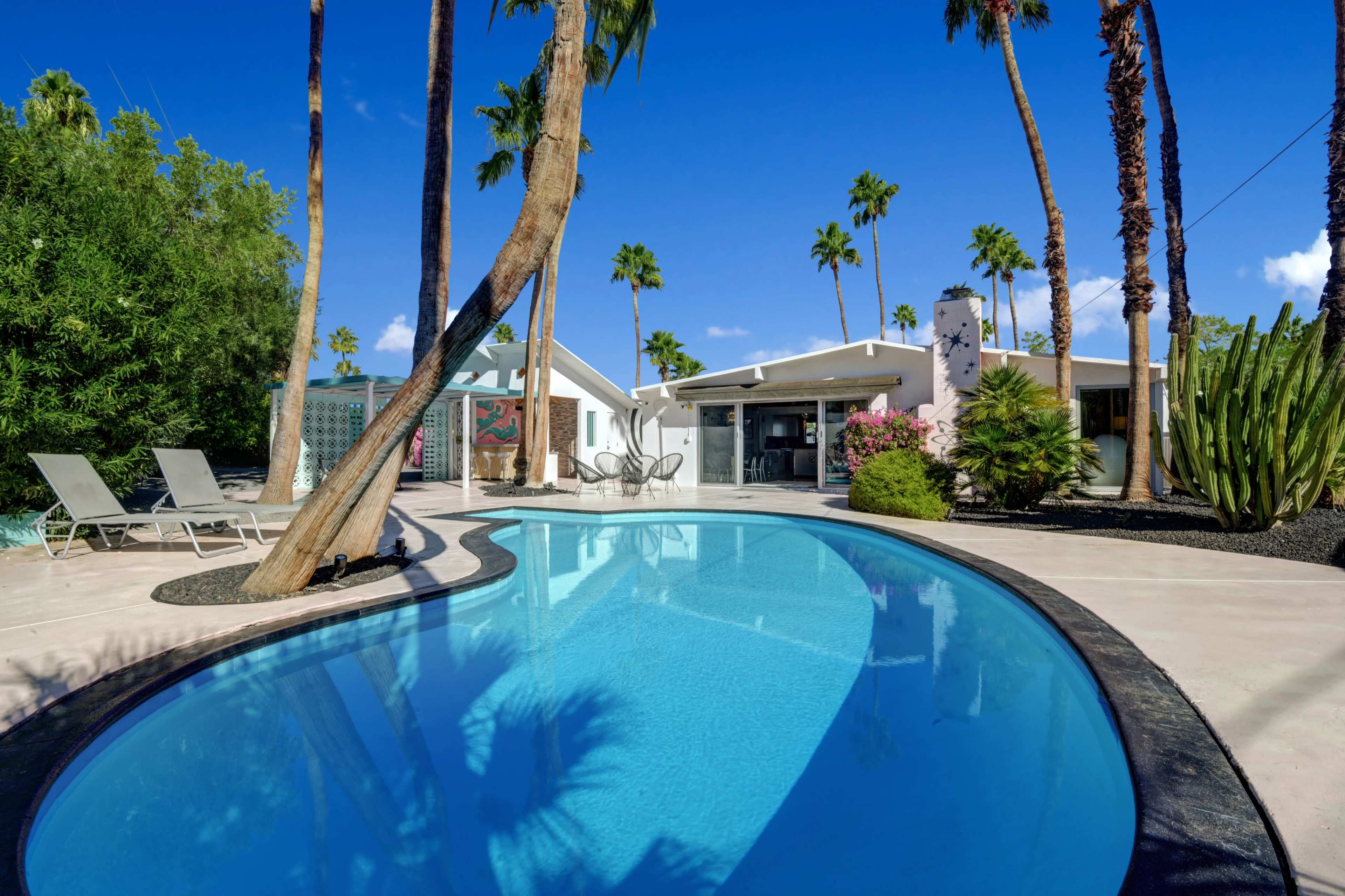 A clear blue swimming pool is surrounded by palm trees and a modern house, with lounge chairs placed nearby.