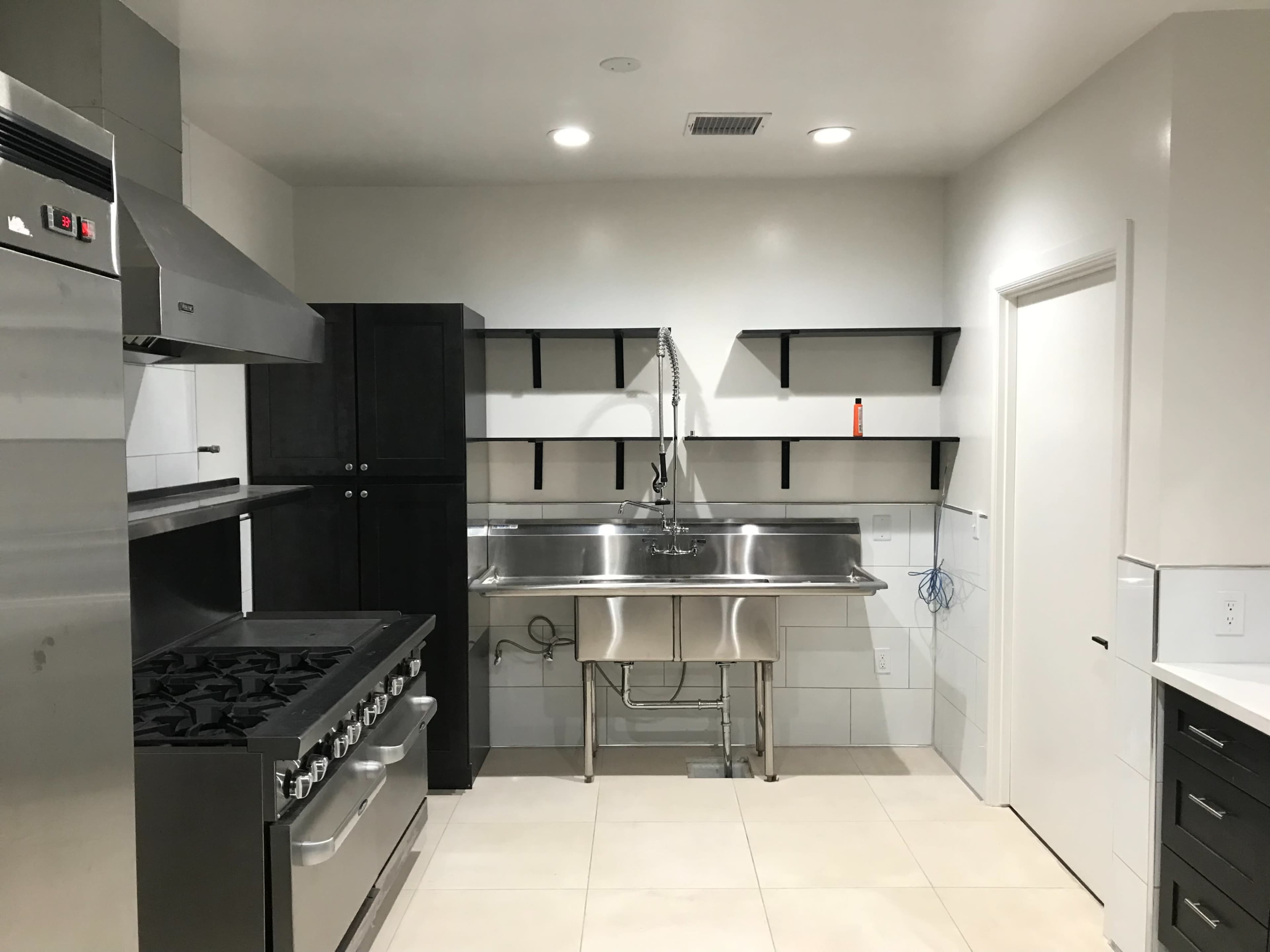 A modern kitchen featuring stainless steel appliances, a large gas stove, a stainless steel sink with open shelves above, and light-colored tiles on the floor.
