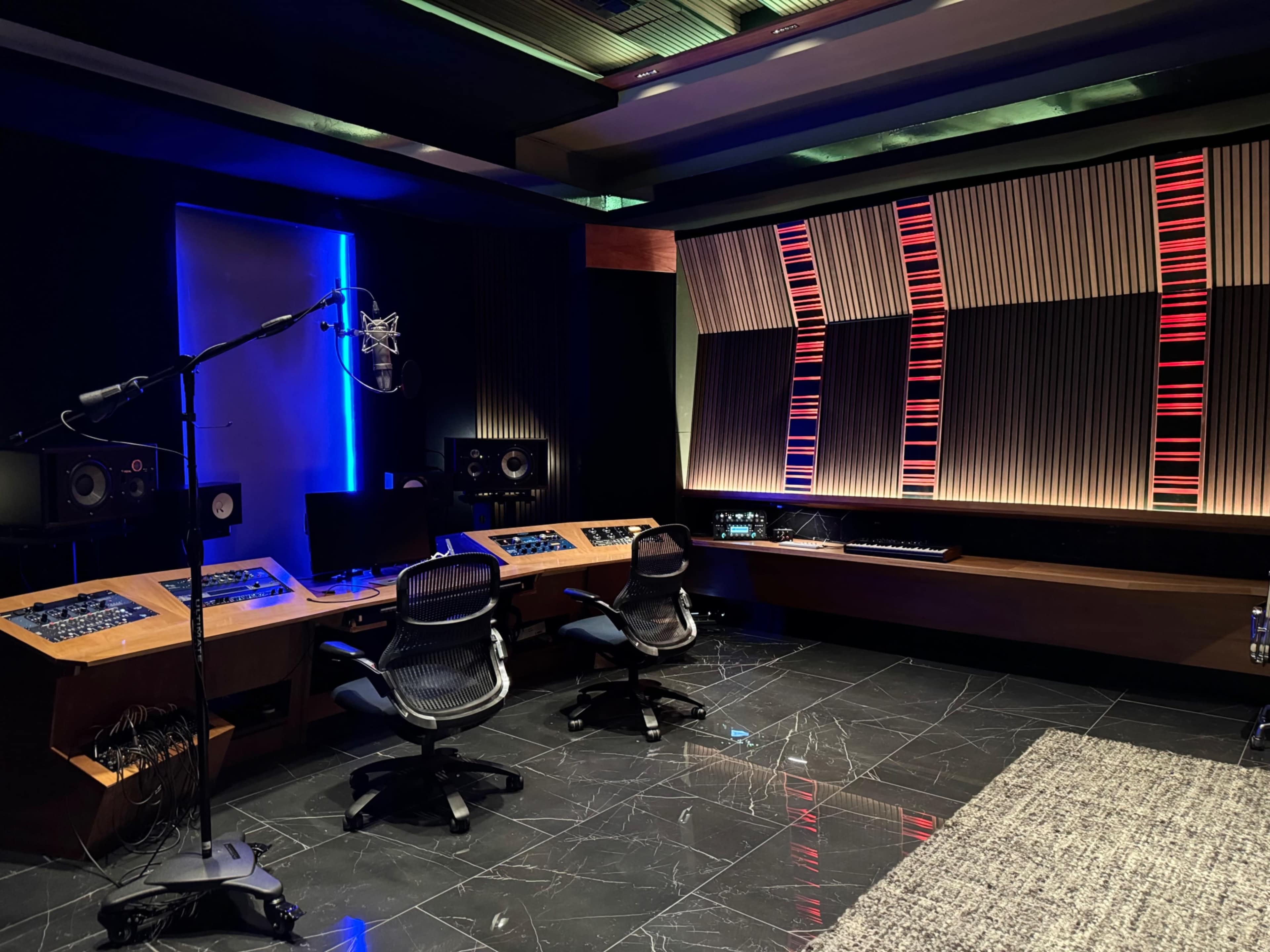 A modern music recording studio featuring a microphone, sound mixing equipment, and two chairs facing a soundboard, with a large screen and illuminated panels on the walls.