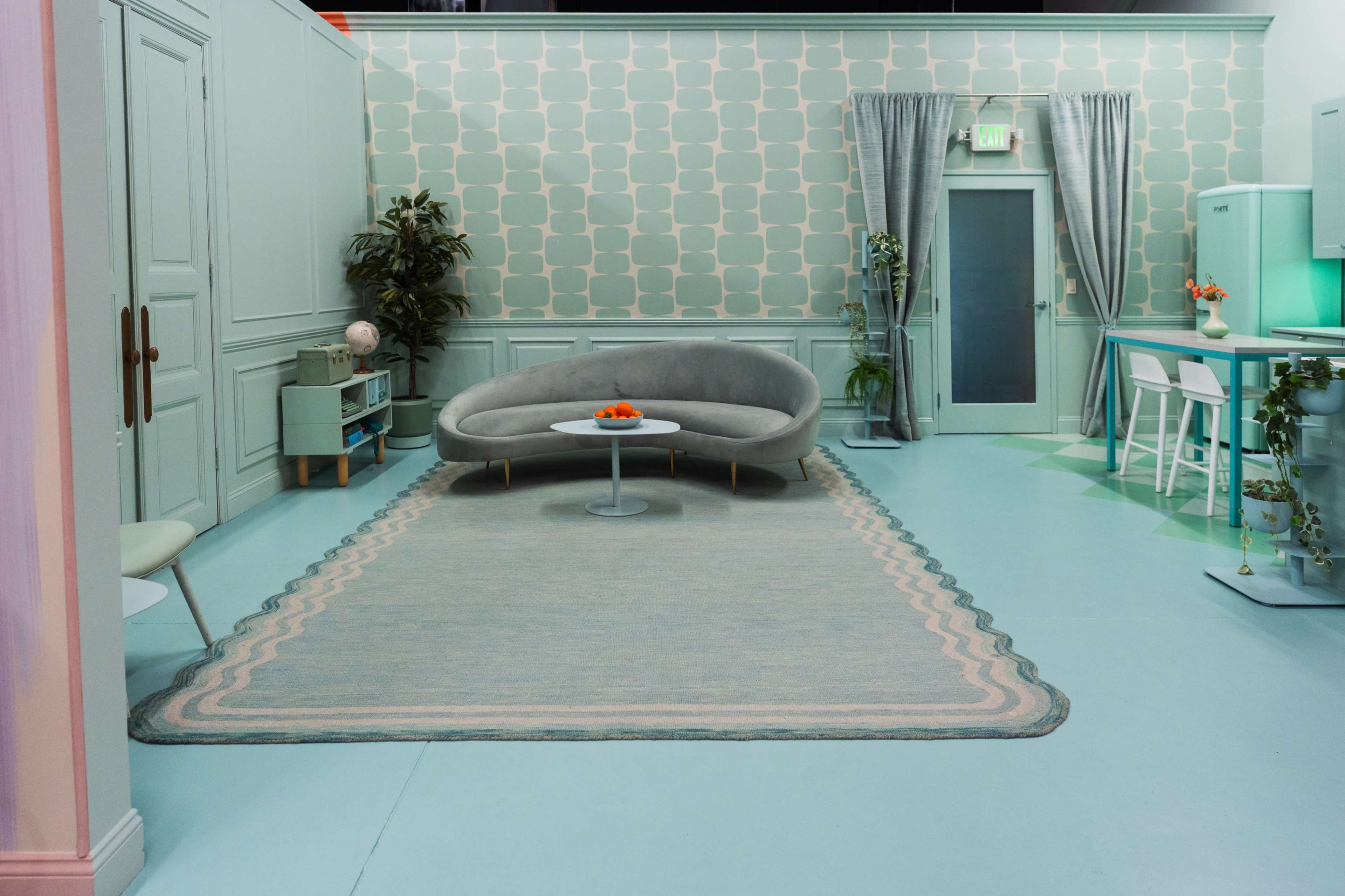 The image shows a modern, minimally furnished room with light green walls, a curved gray sofa, a round table with fruit, and a desk area.