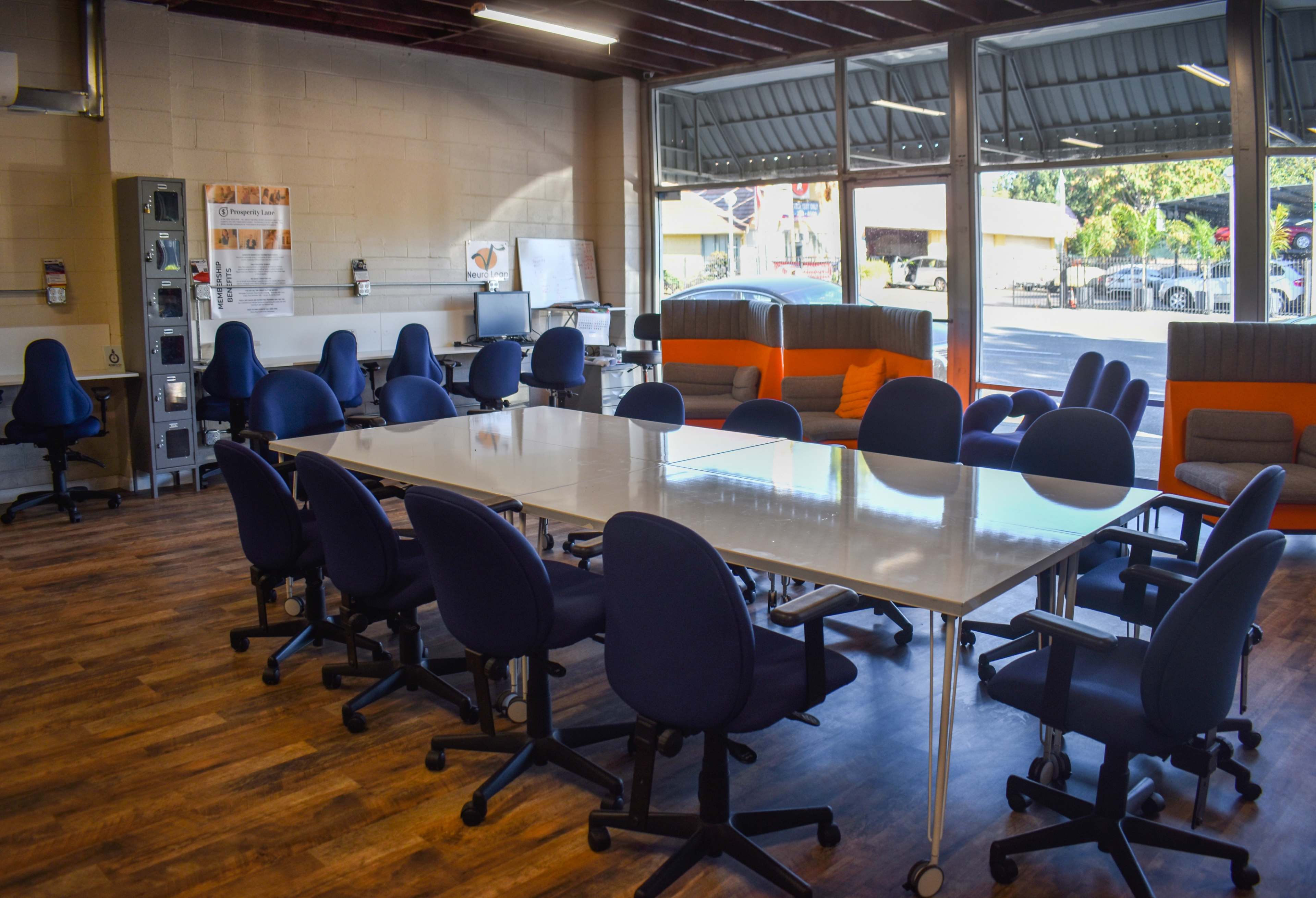 A bright meeting room with a long white table surrounded by blue office chairs, featuring a lounge area with orange and gray seating in the background.