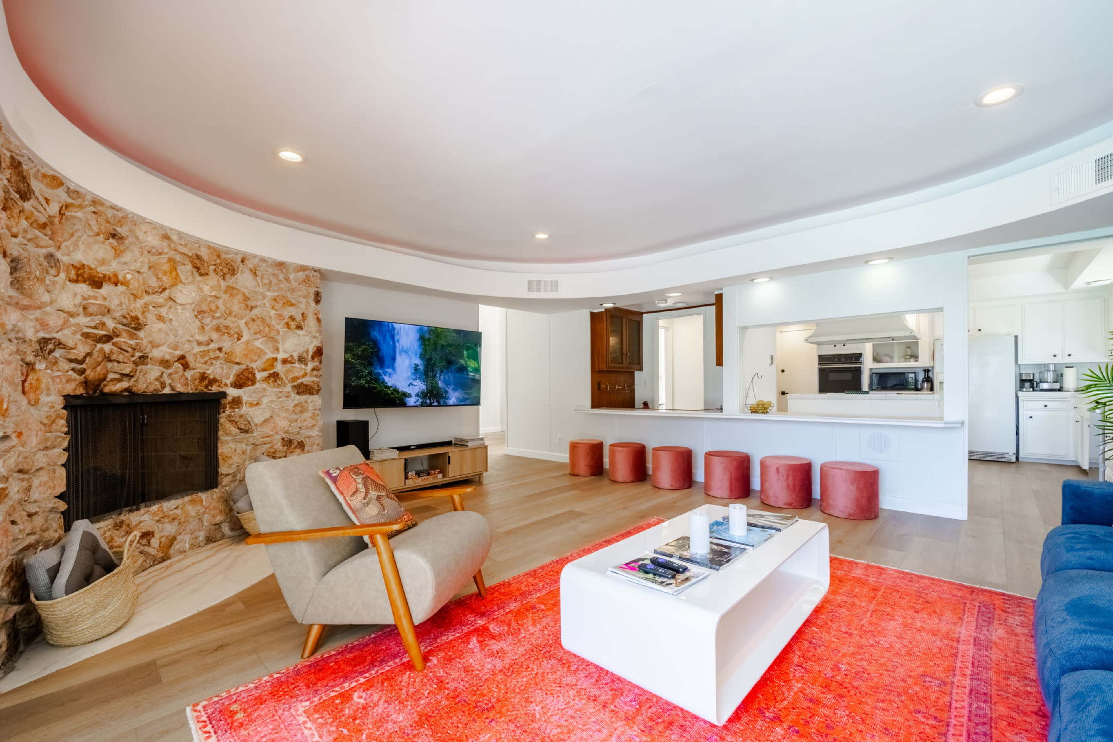 A modern living room featuring a stone fireplace, a large television, and a kitchen area with bar seating.
