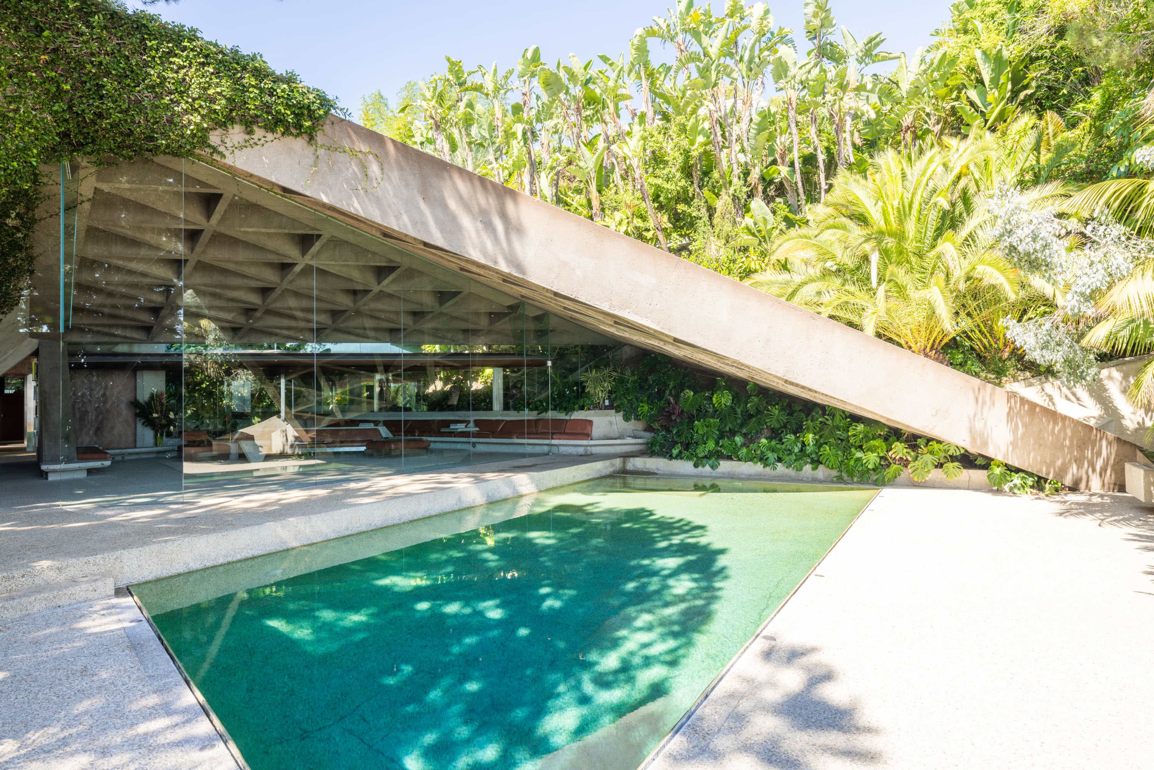 A modern house with a slanted roof, large glass walls, and a swimming pool surrounded by lush tropical plants.