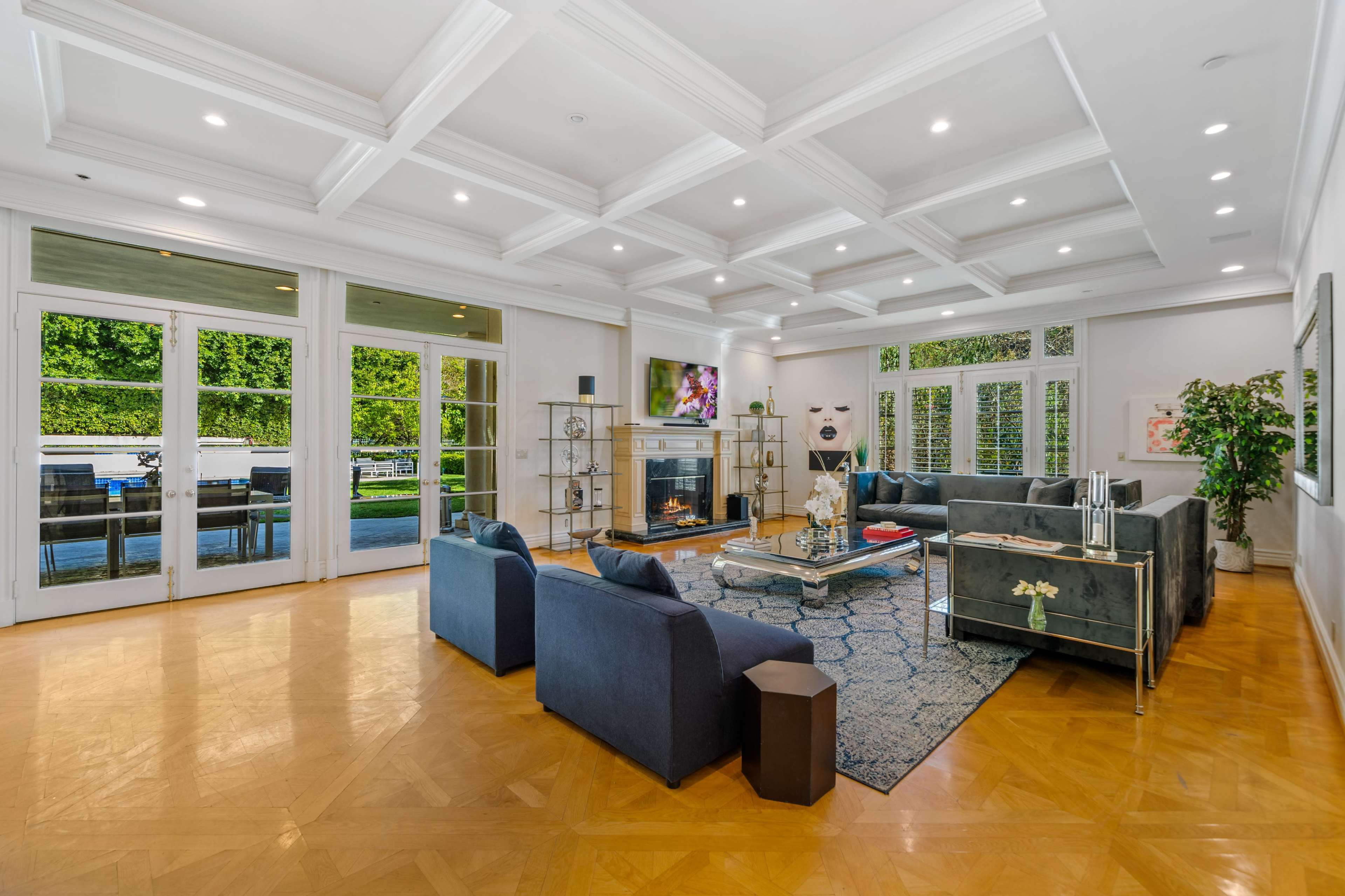 A spacious living room features a coffered ceiling, large windows, and a mix of modern and traditional furnishings.