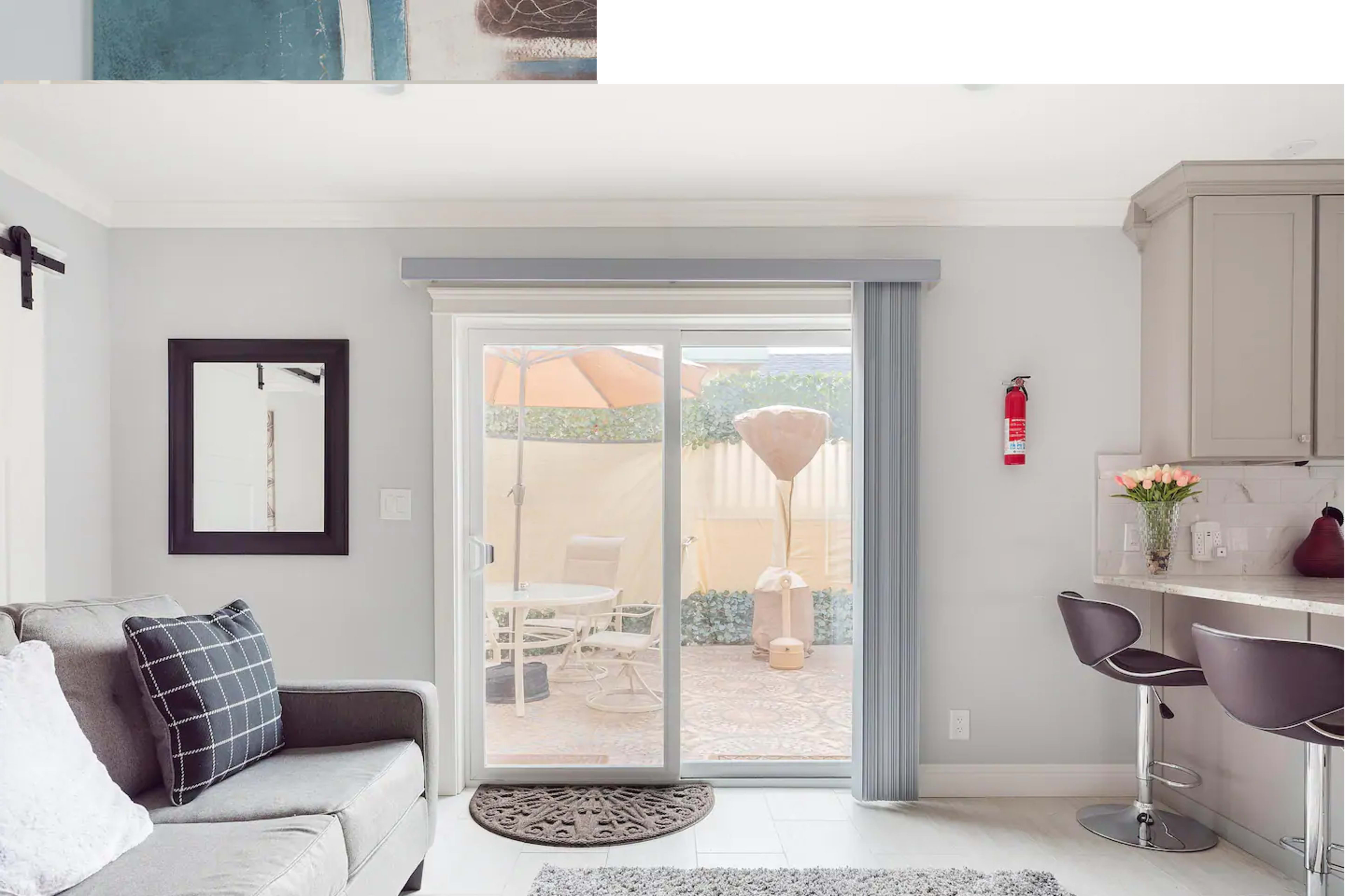 A living space features a gray sofa, a sliding glass door leading to a patio with outdoor furniture, and a kitchen counter with two bar stools.