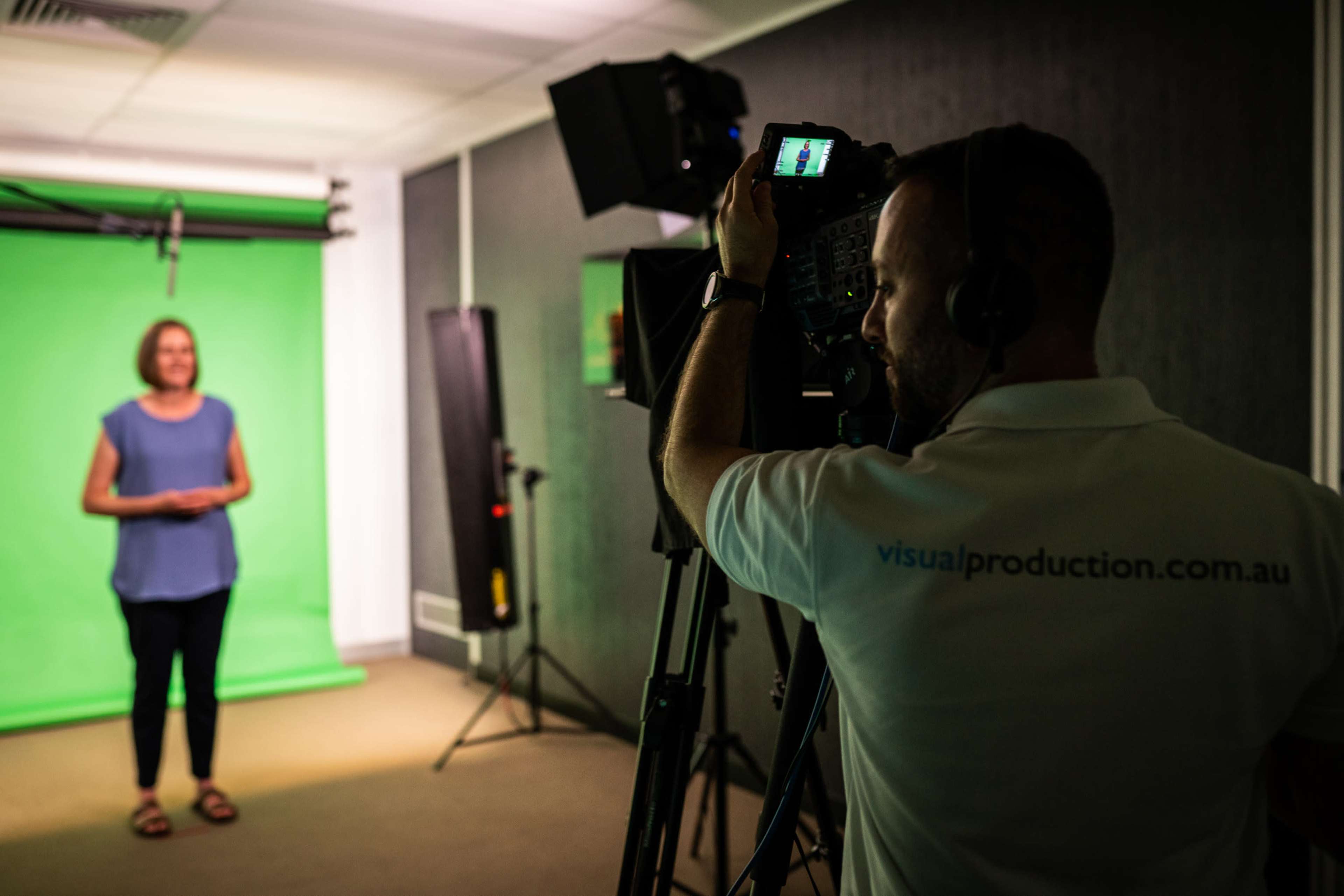 A camera operator filming a woman who is standing in front of a green screen.