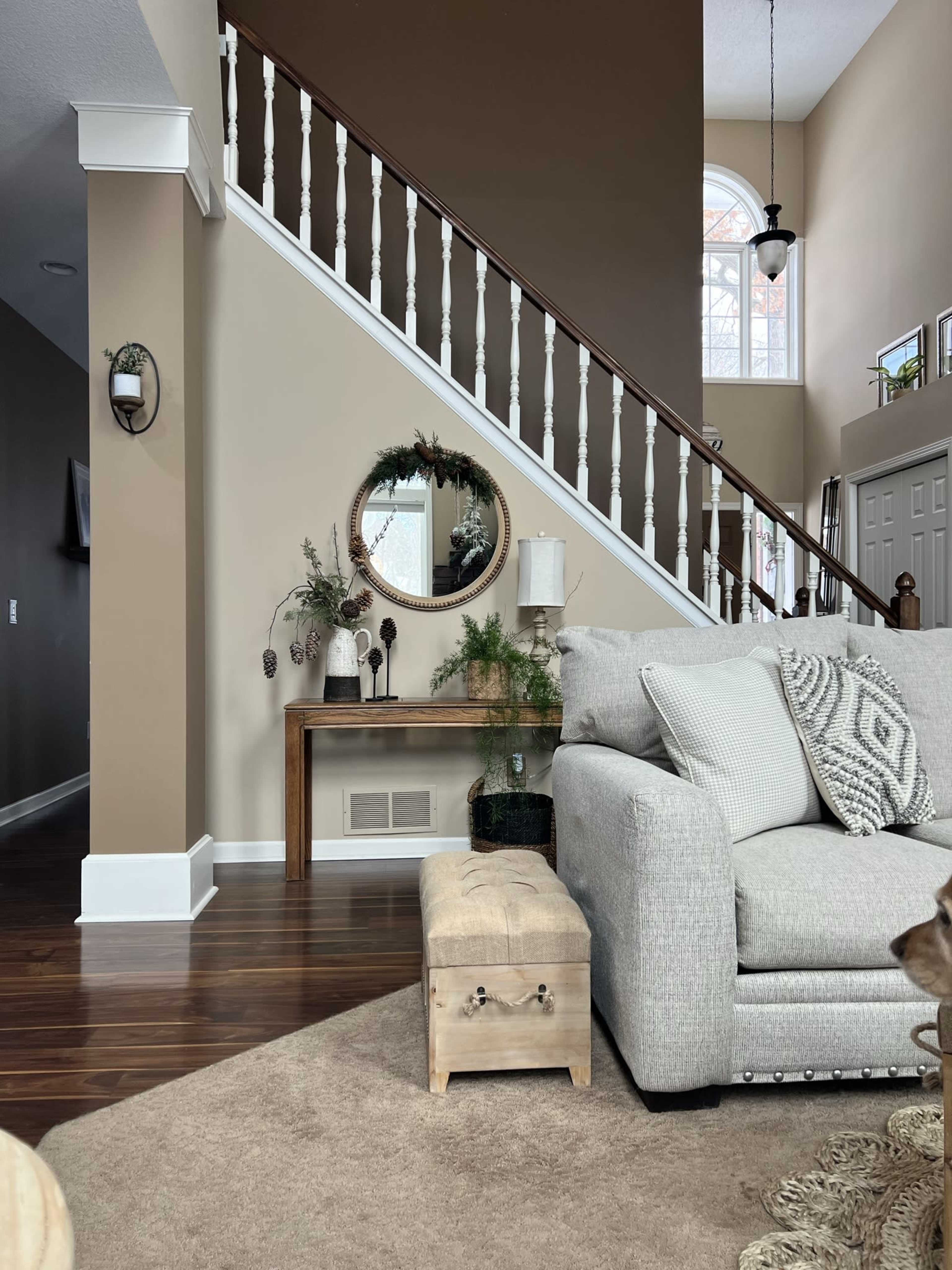 A cozy living room featuring a light gray couch, a wooden bench, and a staircase with white railings, accented by a round mirror on the wall.