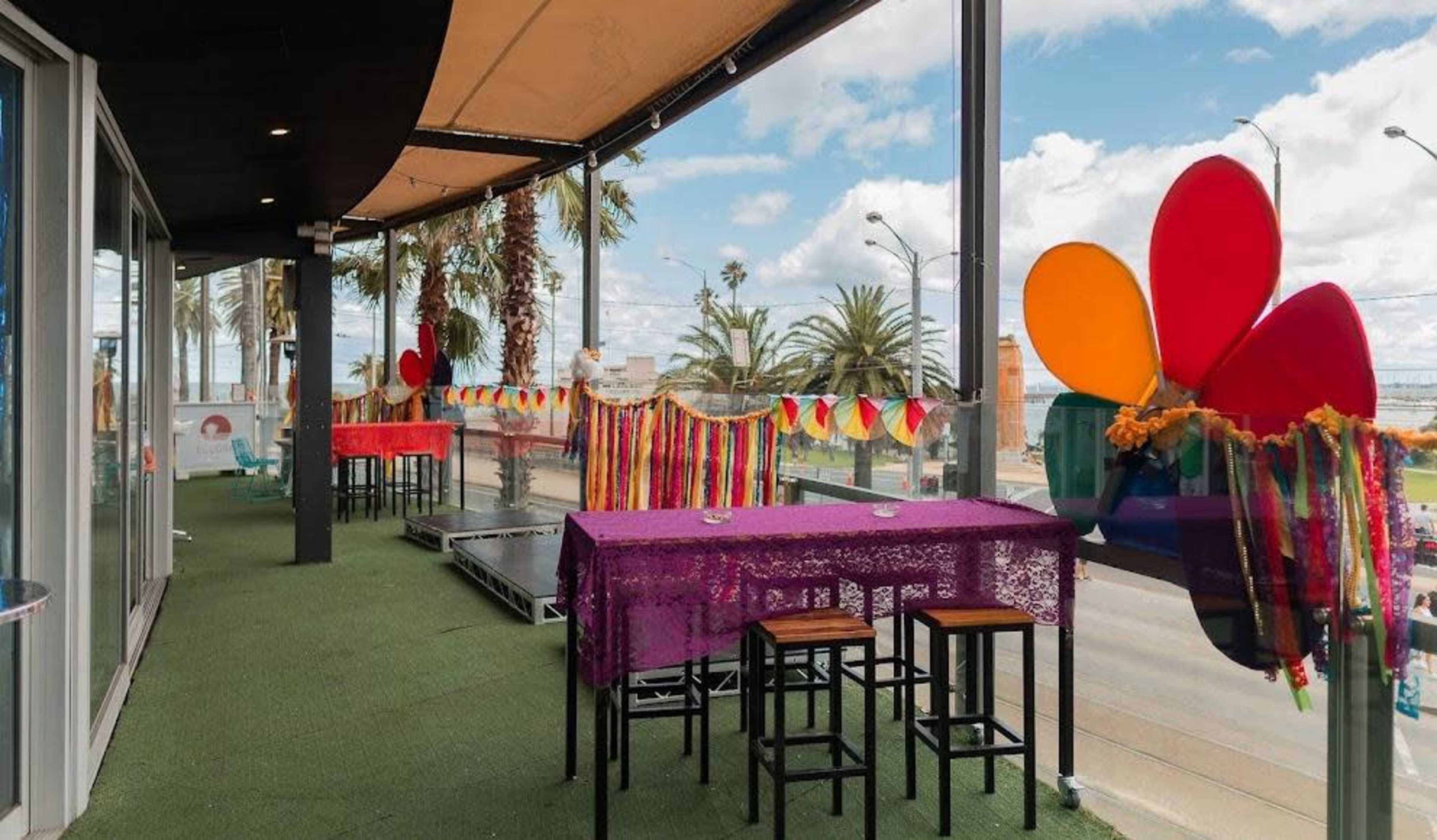 An outdoor terrace with colorful decorations, tables, and chairs overlooking a palm-lined street.