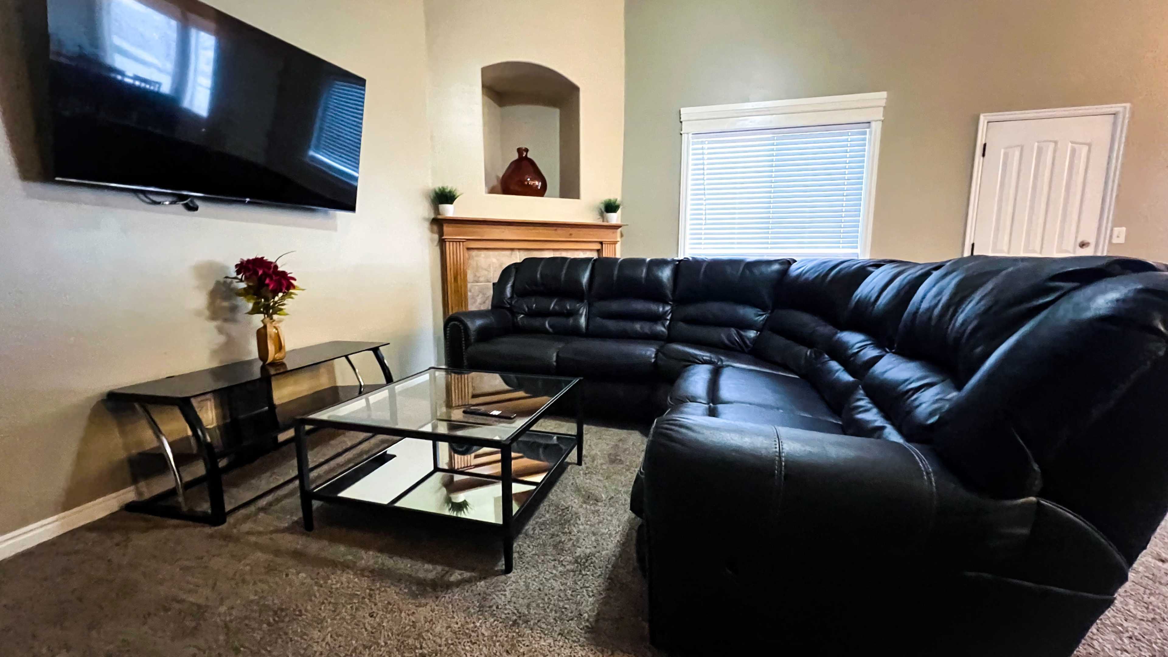 A living room with a black sectional sofa, a glass coffee table, a mounted television, and a window with white blinds.