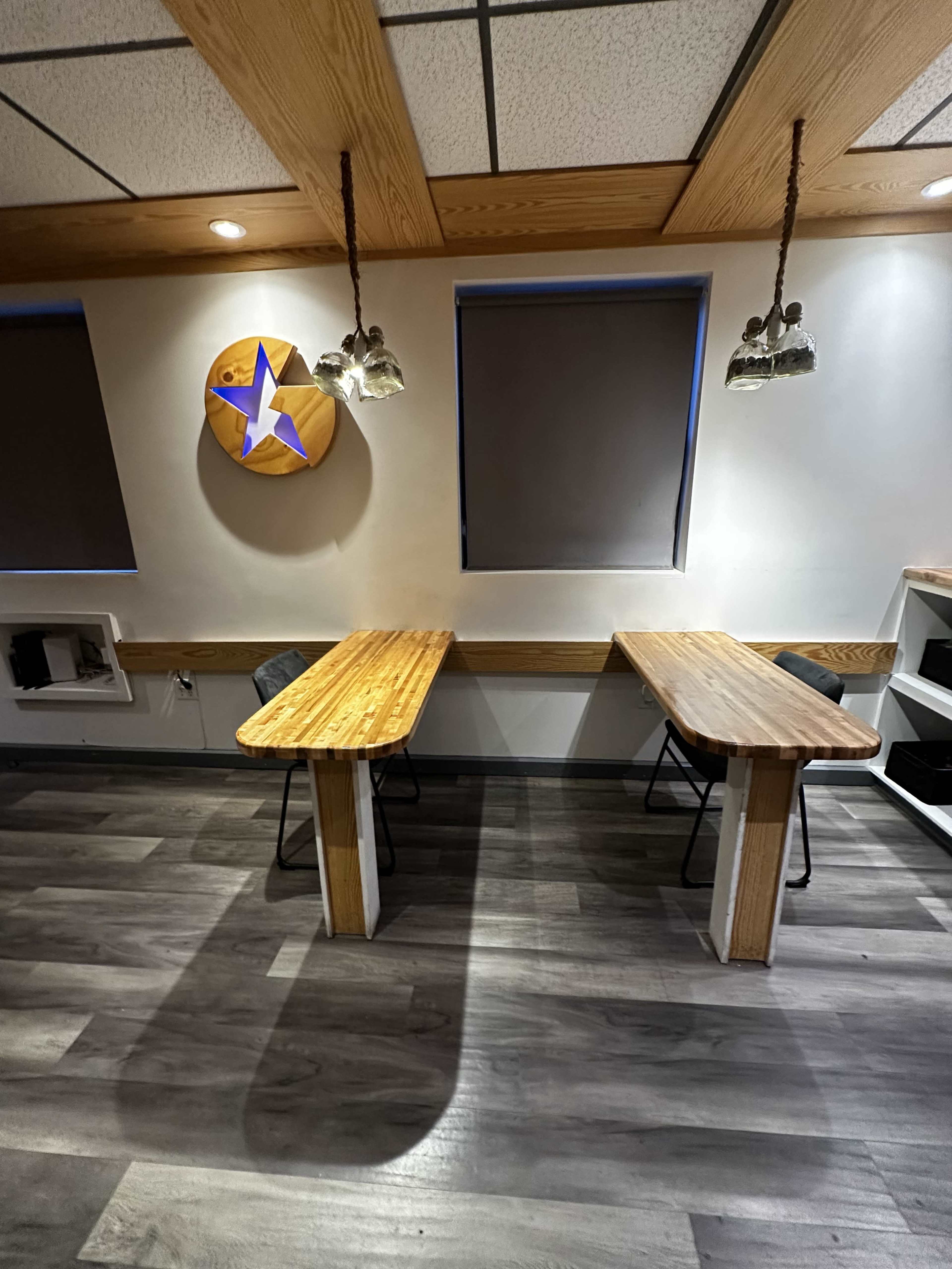 The image shows a room featuring two wooden tables with chairs, gray flooring, and a wall adorned with a circular decorative piece and dim lighting.