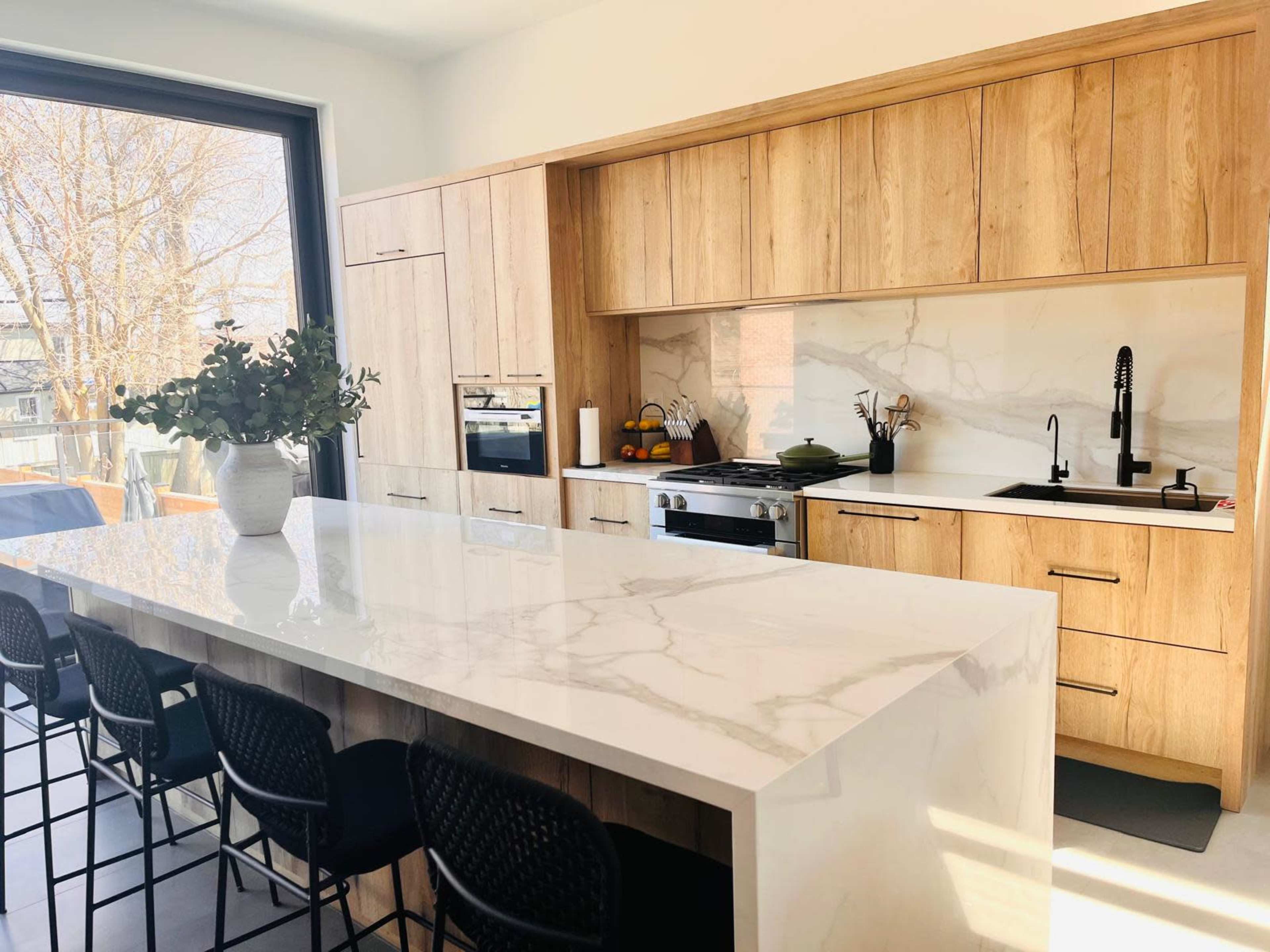 The kitchen features a large white marble island with black bar stools, wooden cabinetry, and a modern stove and sink against a marble backsplash.