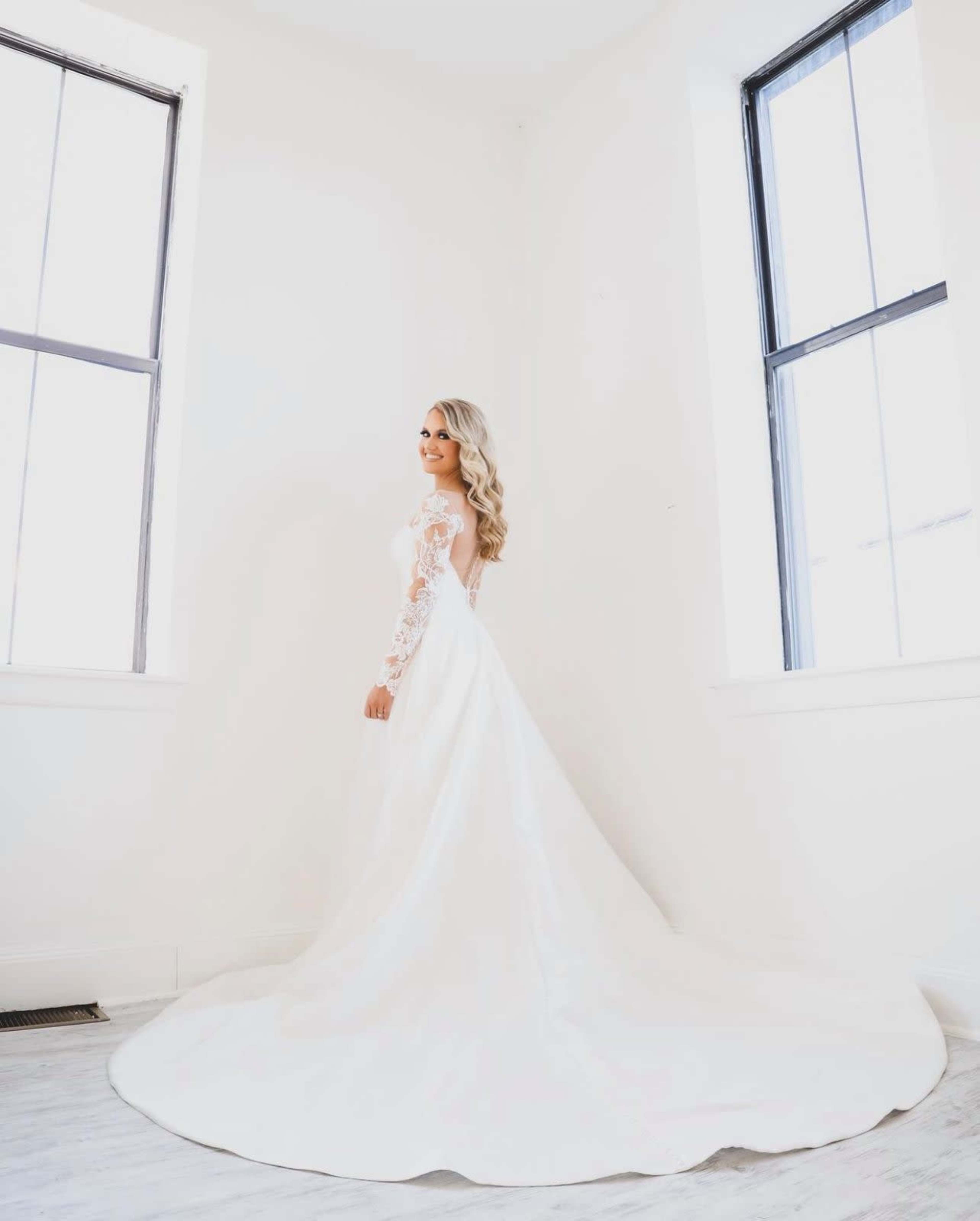 A bride in a white gown with lace sleeves stands gracefully in a well-lit room with two large windows.