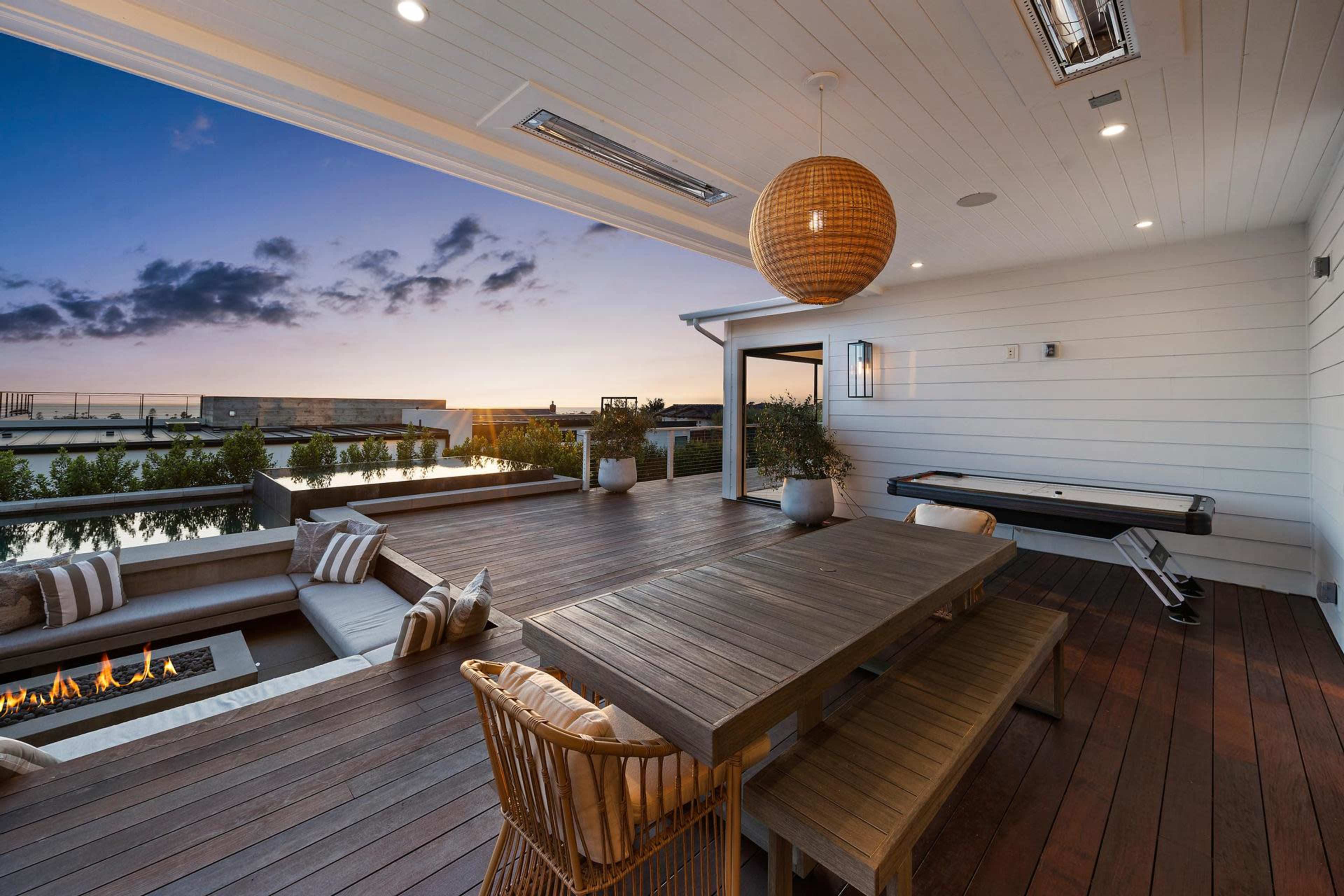The image shows a modern outdoor patio featuring a large wooden dining table, comfortable seating, and a view of the horizon at sunset.