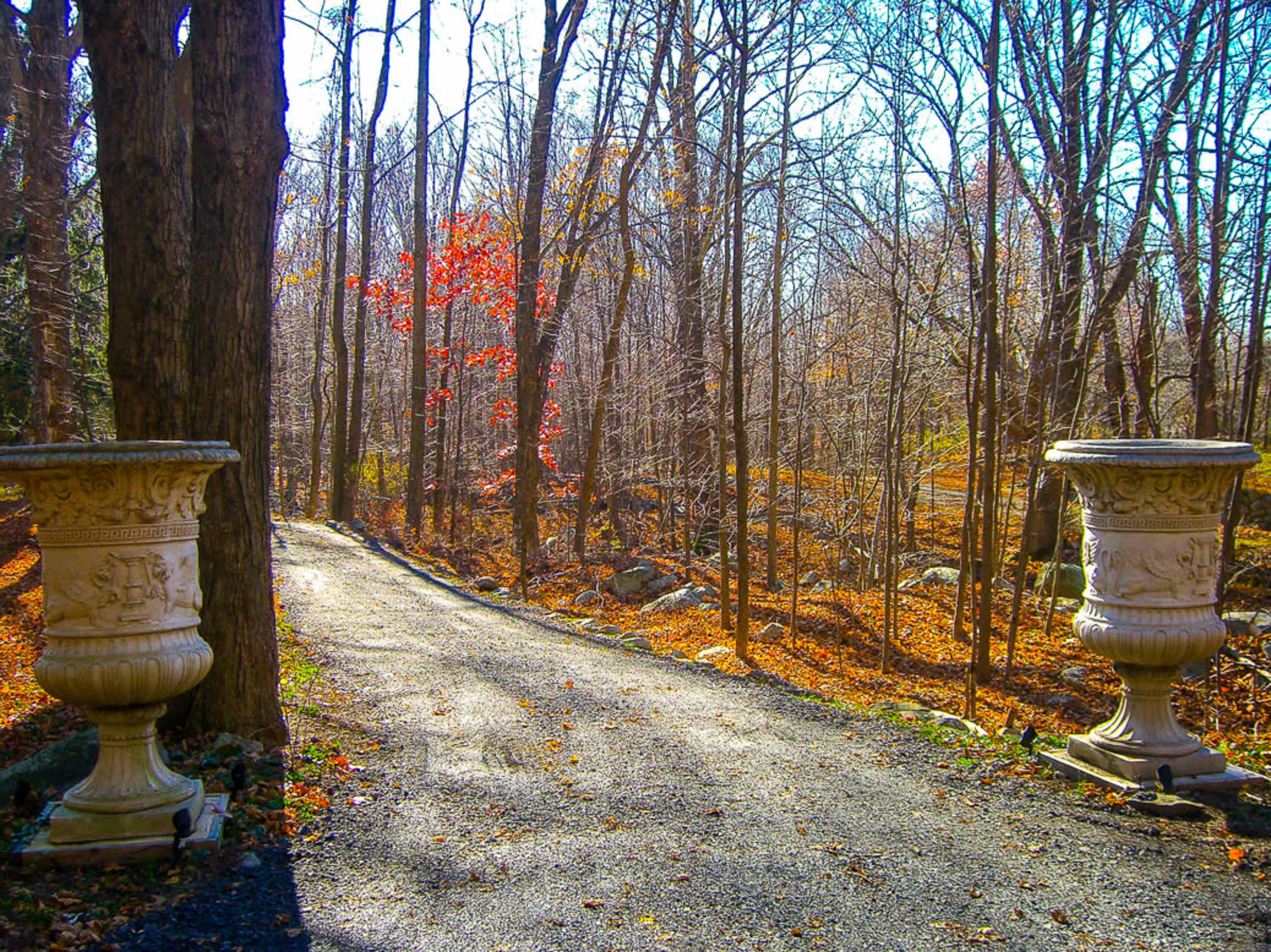 A gravel path winds through a wooded area, flanked by large stone planters, with sparse trees and fallen leaves scattered on the ground.