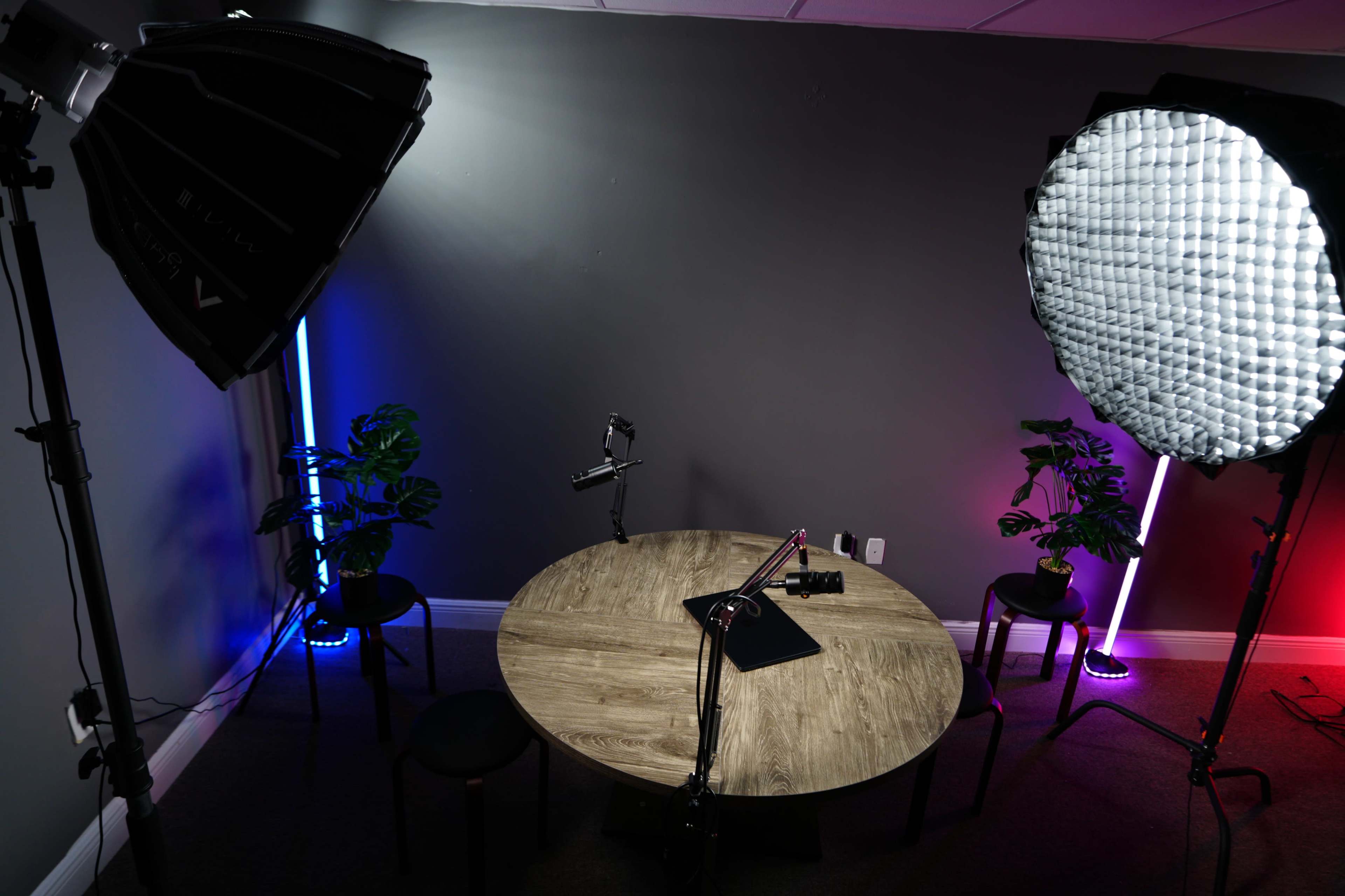The image shows a photography or video setup featuring a round wooden table with a camera and microphone, surrounded by various lighting equipment and two chairs.
